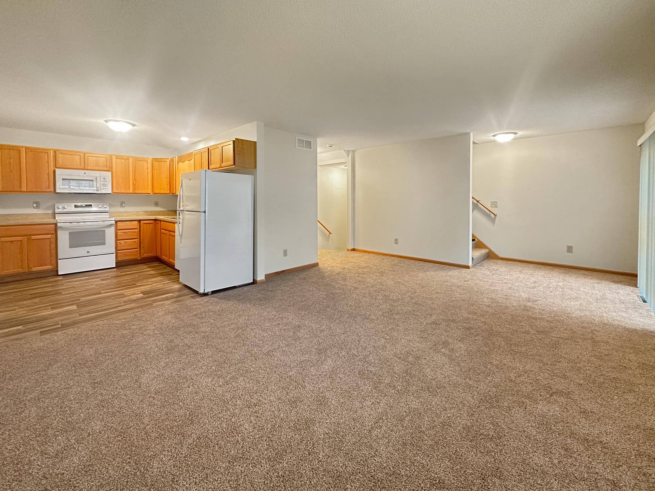 Spacious view of a living area and kitchen in an apartment. The kitchen features wooden cabinets, a white refrigerator, and stove. The living area is carpeted with natural light coming from a window, and there’s a staircase leading to another level in the background. Ceiling lights provide illumination.