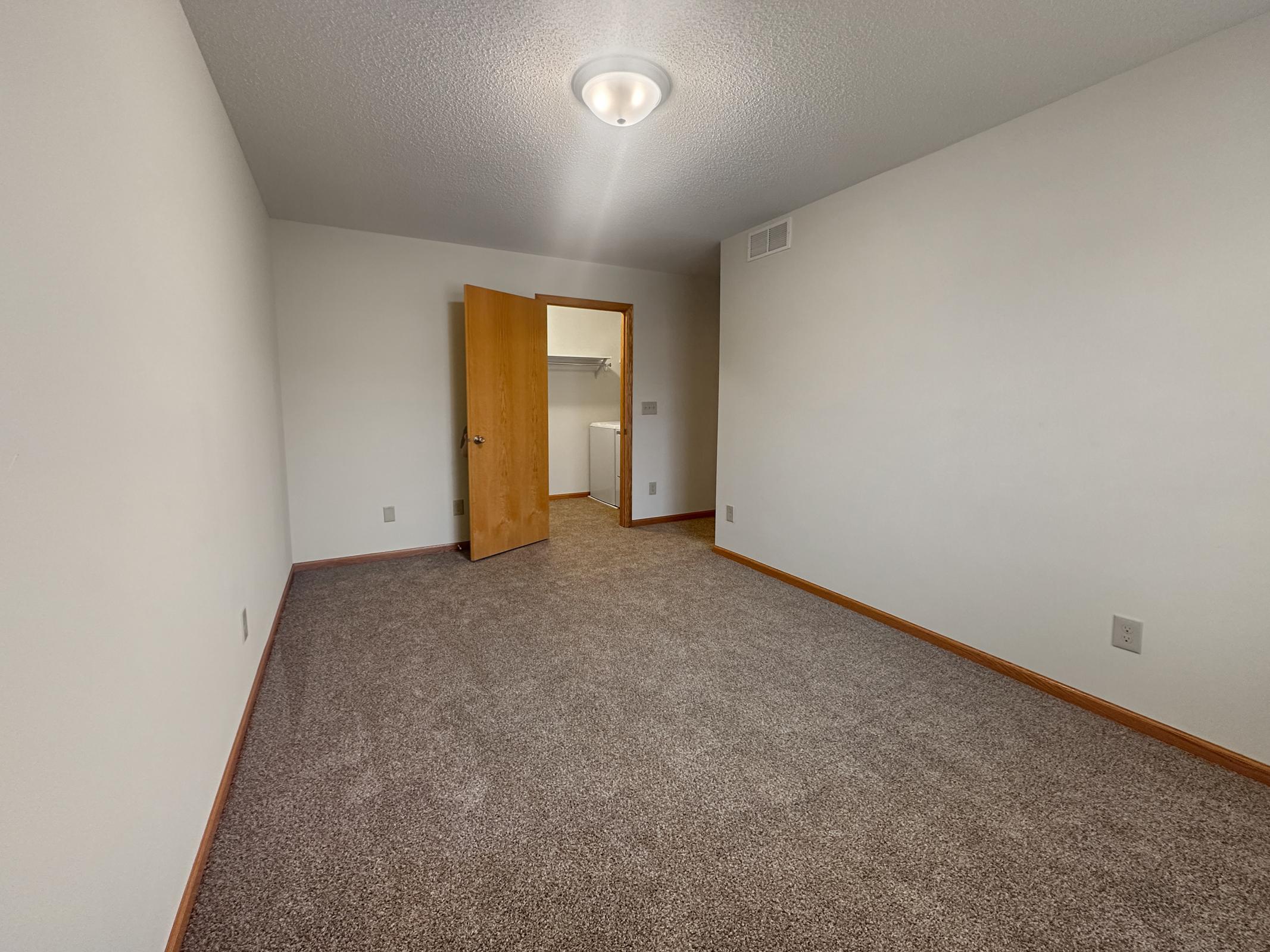 A vacant room with light-colored walls and a carpeted floor. There is a light fixture on the ceiling, and a wooden door leads to another space. The room is empty, providing a spacious feel with no furniture present. Natural light may enter through the door area.