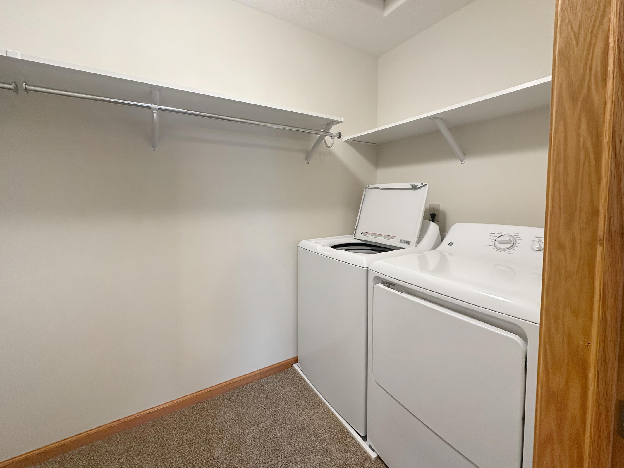 A clean and organized laundry room featuring a white top-loading washing machine and a white dryer. There is a shelf above the appliances for storage, and the walls are painted a light color. The floor is carpeted, providing a cozy feel to the space.