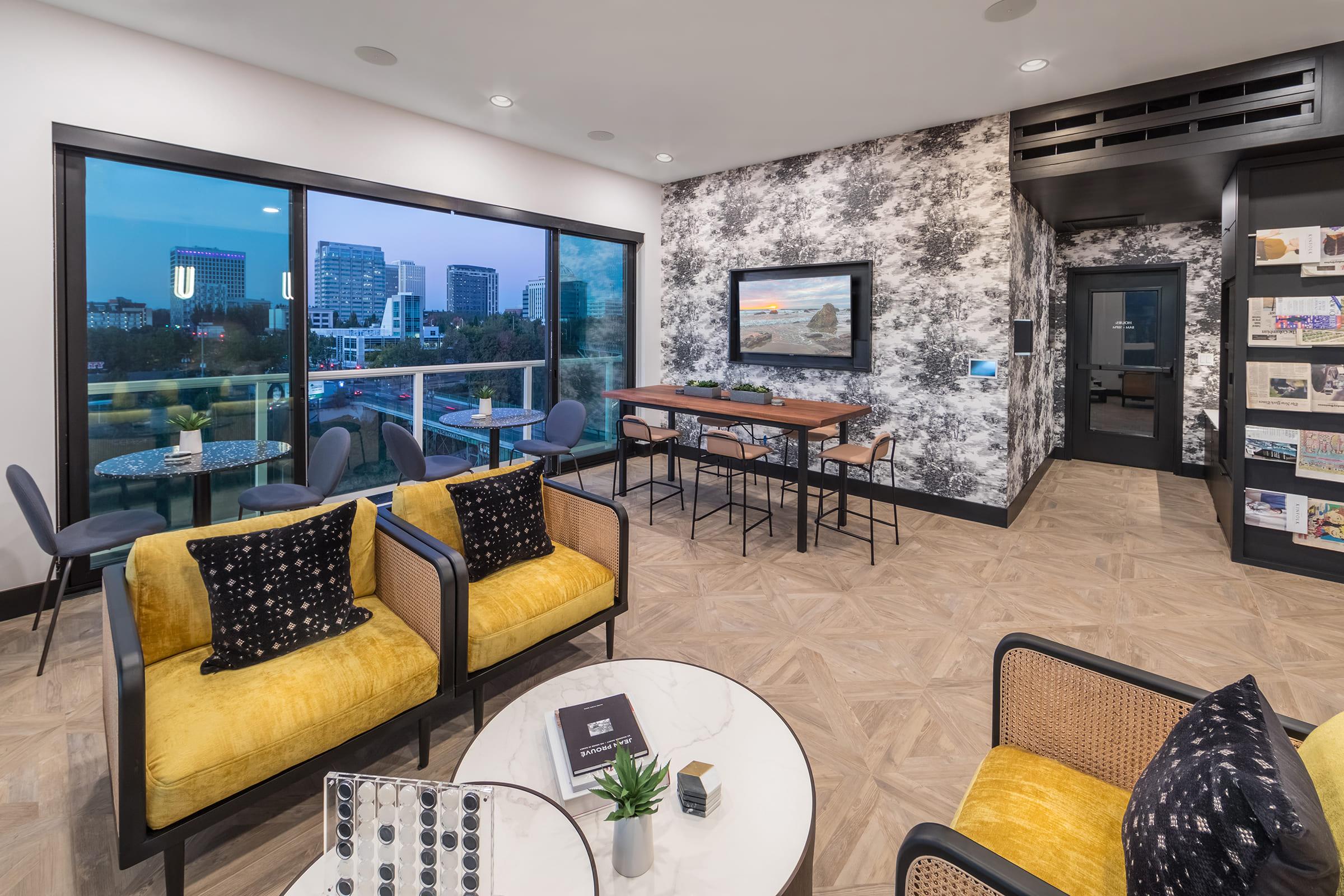 A modern lounge area featuring stylish yellow armchairs and a table. Large windows offer a view of a cityscape at dusk. A textured wall adds character, while a large painting and shelves with decorative items enhance the decor. The space is well-lit and inviting, ideal for relaxation or social gatherings.