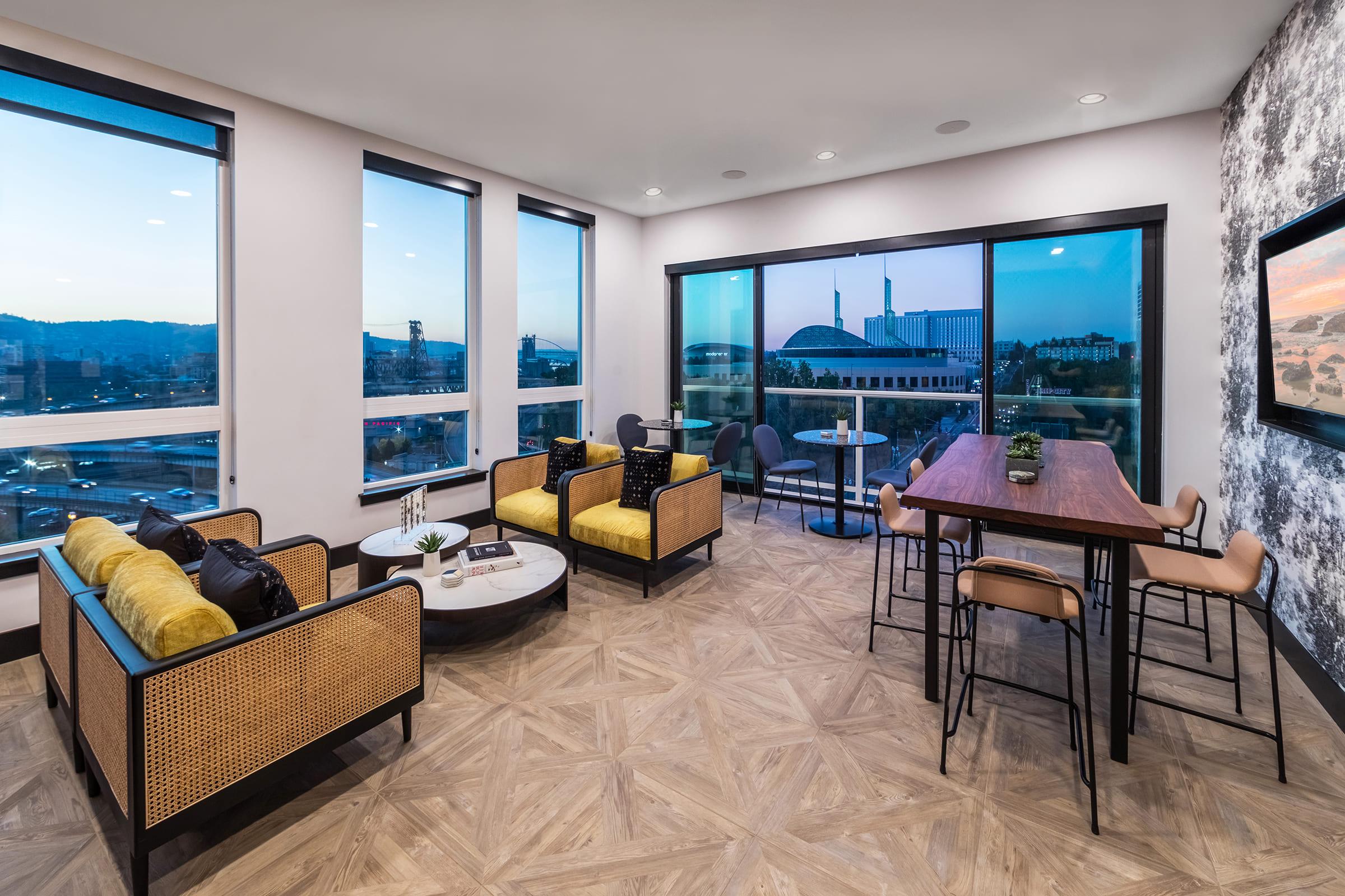 Interior view of a modern living space featuring a cozy seating area with yellow and black upholstered chairs, a round coffee table, and a dining table with chairs. Large windows offer a panoramic view of the city skyline at twilight, with soft lighting enhancing the contemporary design.