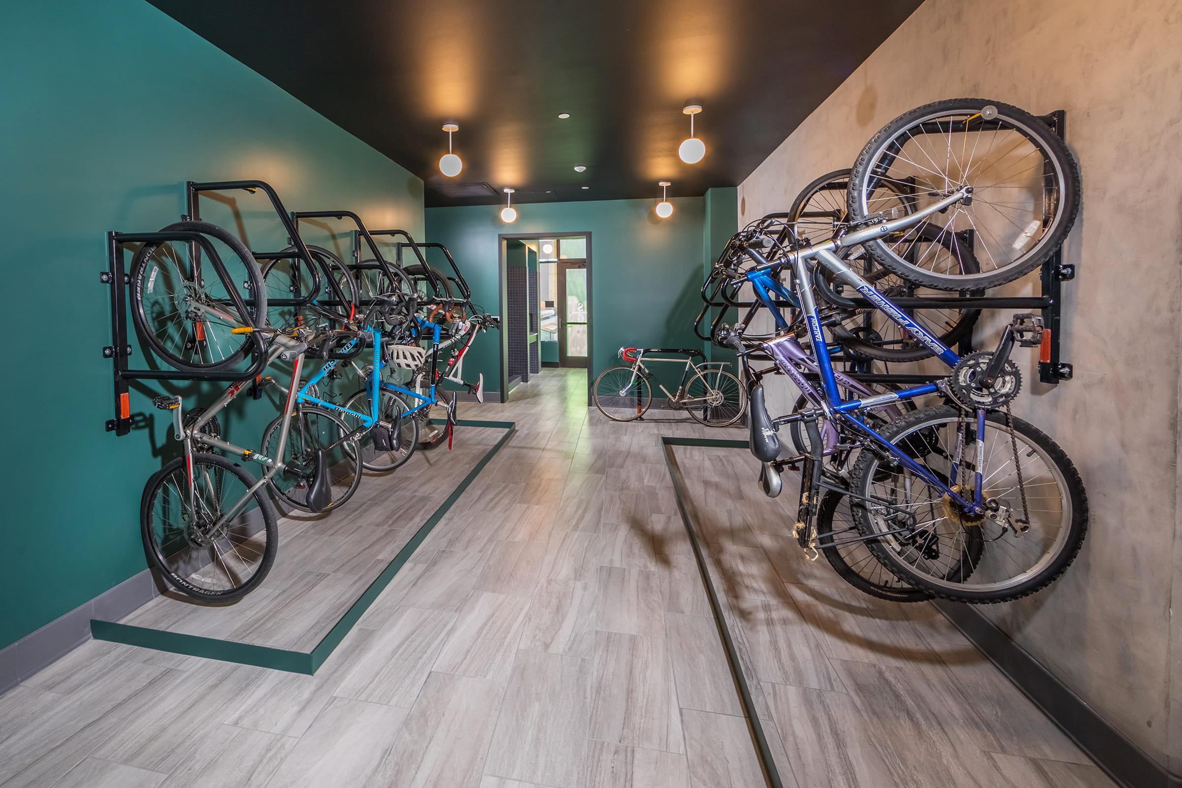 A modern bike storage area featuring multiple bicycle racks mounted on green walls. The space includes various types of bicycles, with some hanging and others resting on the floor. The flooring is light-colored tiles, and the lighting is provided by stylish hanging bulbs, creating a contemporary atmosphere.