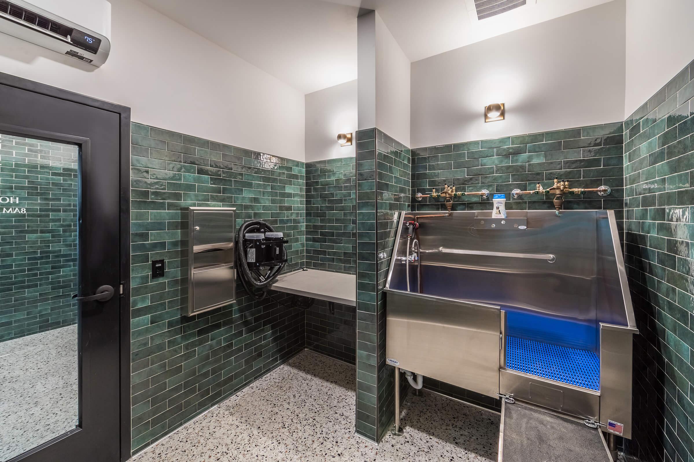 A modern pet washing station featuring green tile walls, a stainless steel bath with a blue water feature, and a large sink. The area is well-lit with wall sconces and includes a space for pet grooming equipment, designed for convenience and cleanliness.