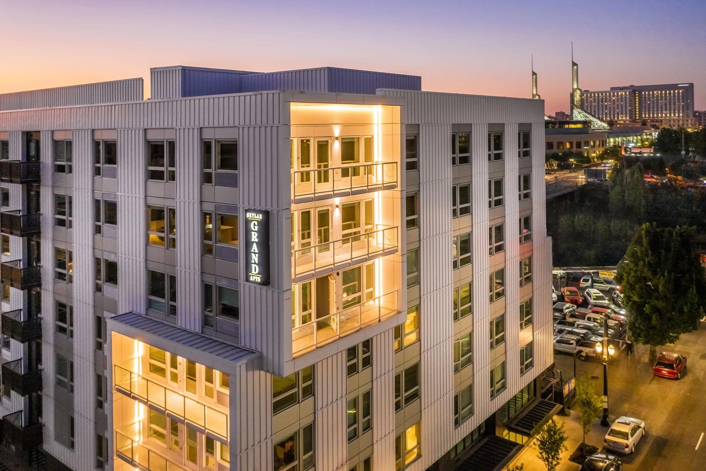 A modern multi-story building with illuminated balconies at dusk, featuring a sign that reads "The Grand." The surrounding area includes parked cars and a view of city buildings in the background, showcasing a tranquil urban environment at sunset.