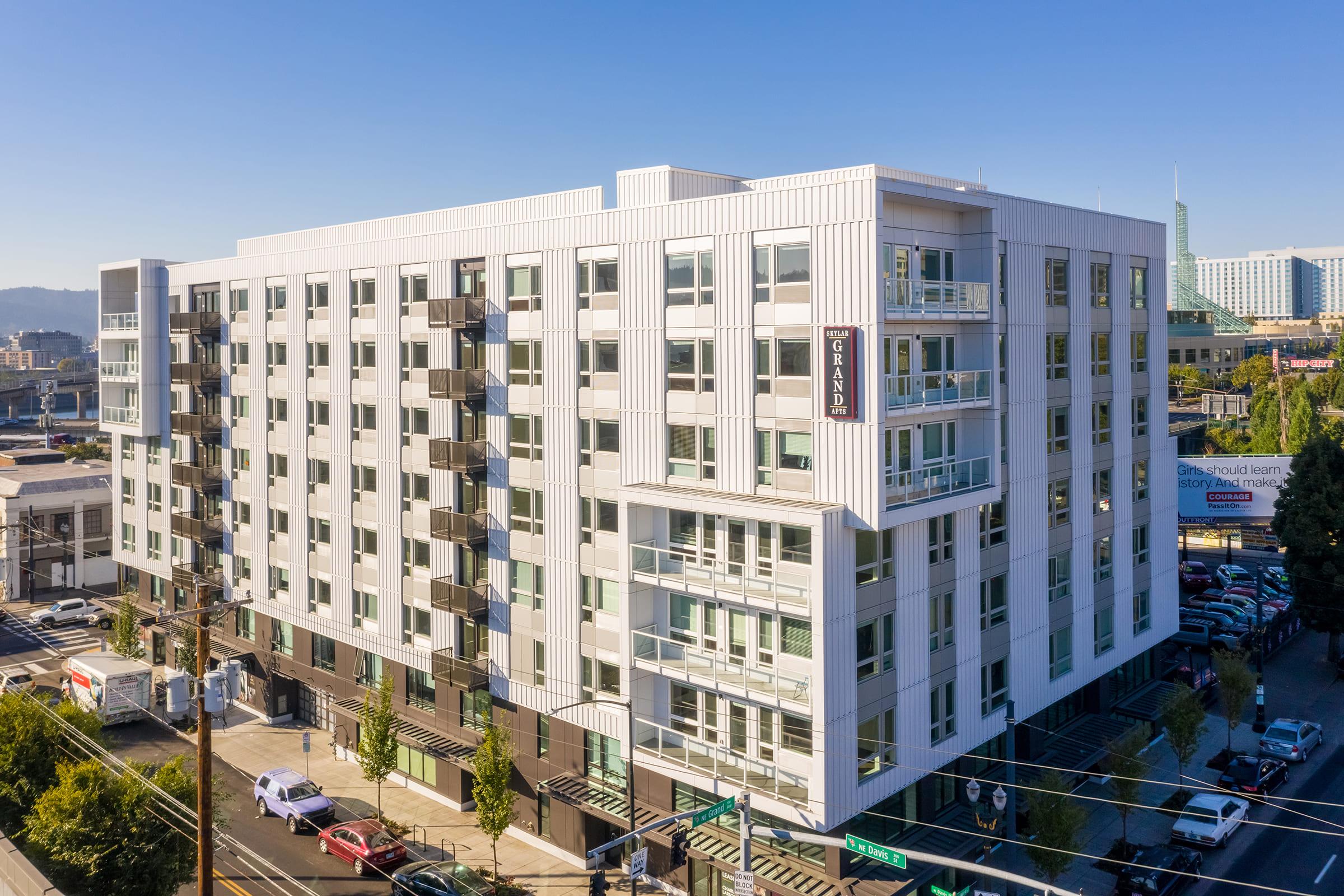 A modern, multi-story apartment building with a sleek white exterior. It features balconies, large windows, and is situated in an urban environment with trees and parked cars nearby. The sky is clear and blue, highlighting the building's contemporary design.