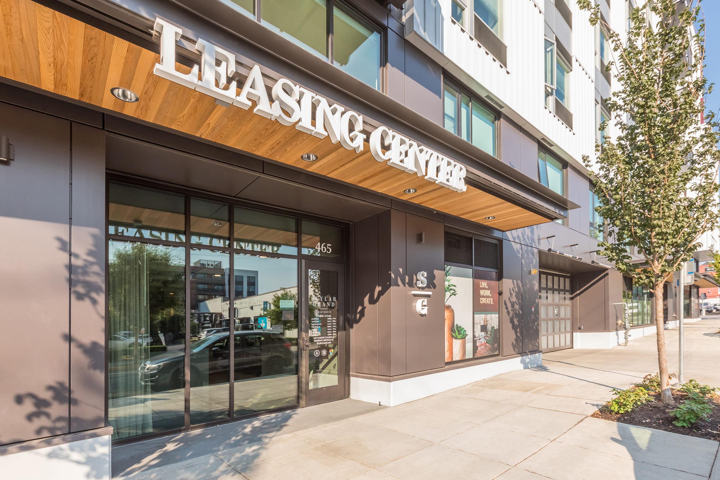 Exterior view of a modern leasing center with large windows and a wooden overhang. The building features a sleek design, with a sign reading "Leasing Center" prominently displayed above the entrance. Surrounding area includes landscaped greenery and a sidewalk.
