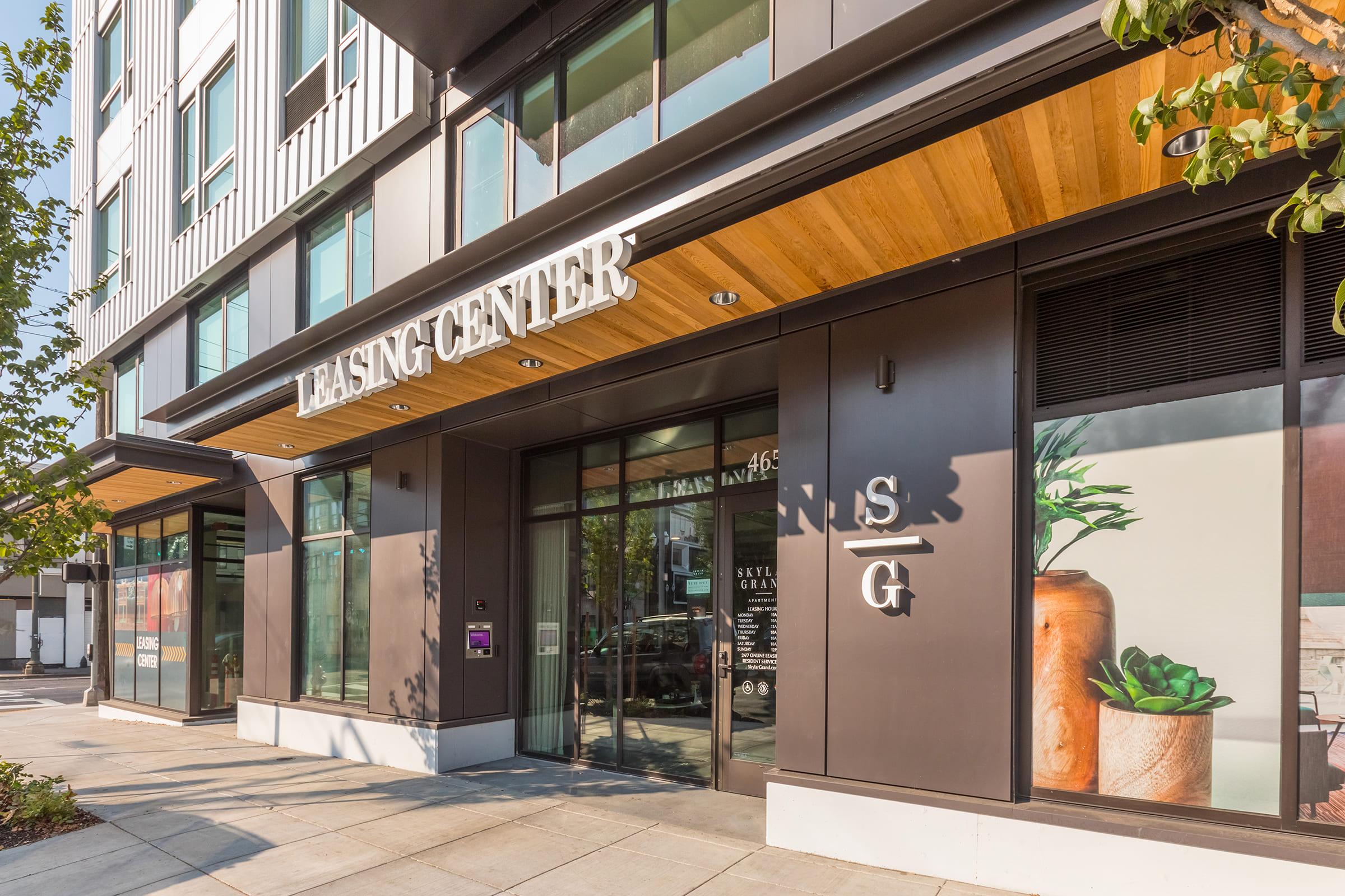 Exterior view of a modern leasing center with large glass windows and a wooden awning. The building features a clean design with prominent signage that reads "Leasing Center." Surrounding greenery enhances the contemporary urban setting.