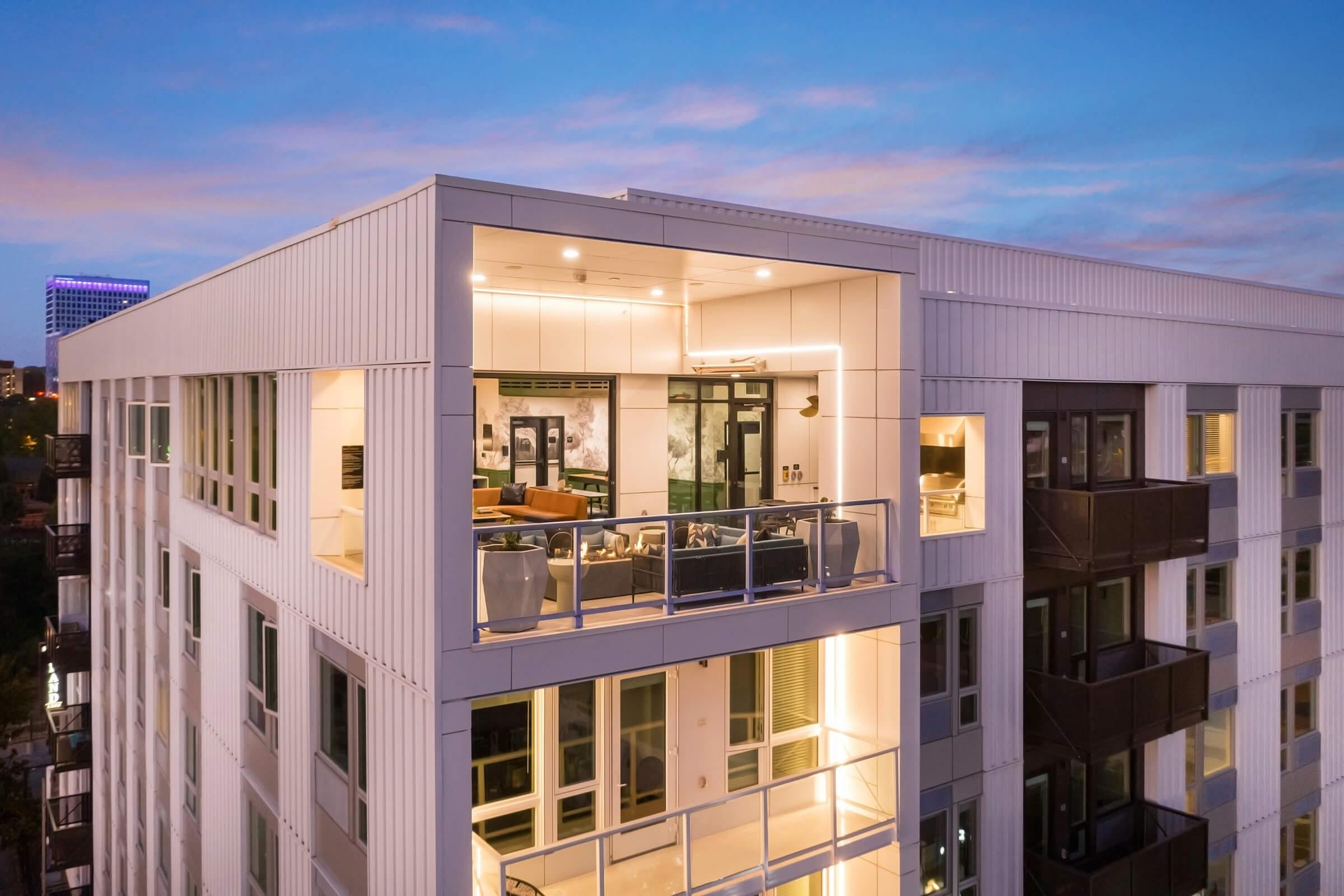 Modern apartment building with a sleek design, featuring large glass windows and a spacious balcony. Interior lights are on, illuminating a stylish living space. The scene captures a sunset sky, highlighting the building's contemporary architecture in an urban setting.