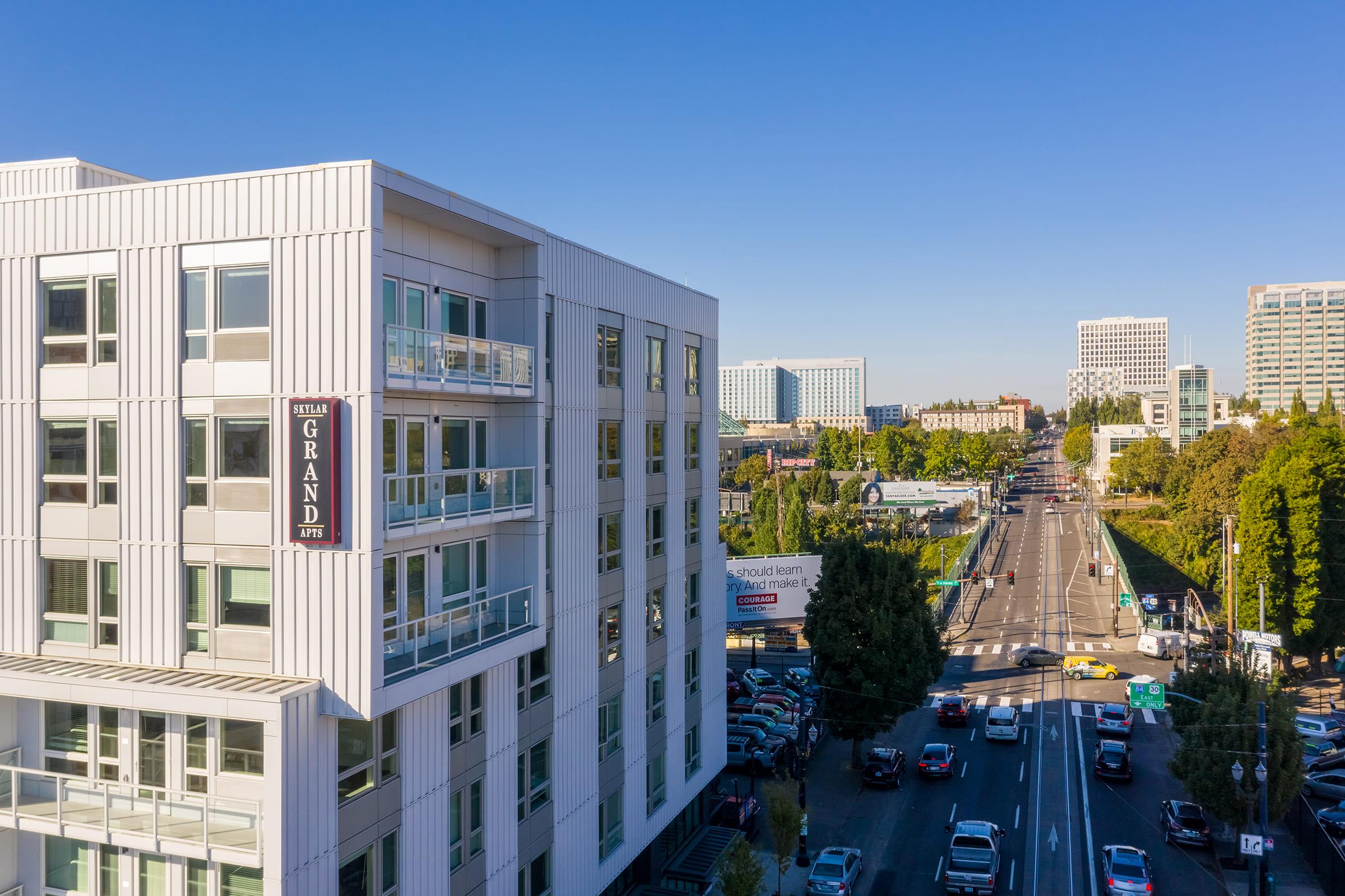 A modern multi-story building with the sign "GRAND" visible, overlooking a city street lined with trees and parked cars. In the background, various commercial buildings and a clear blue sky are present, depicting an urban landscape with a vibrant atmosphere.