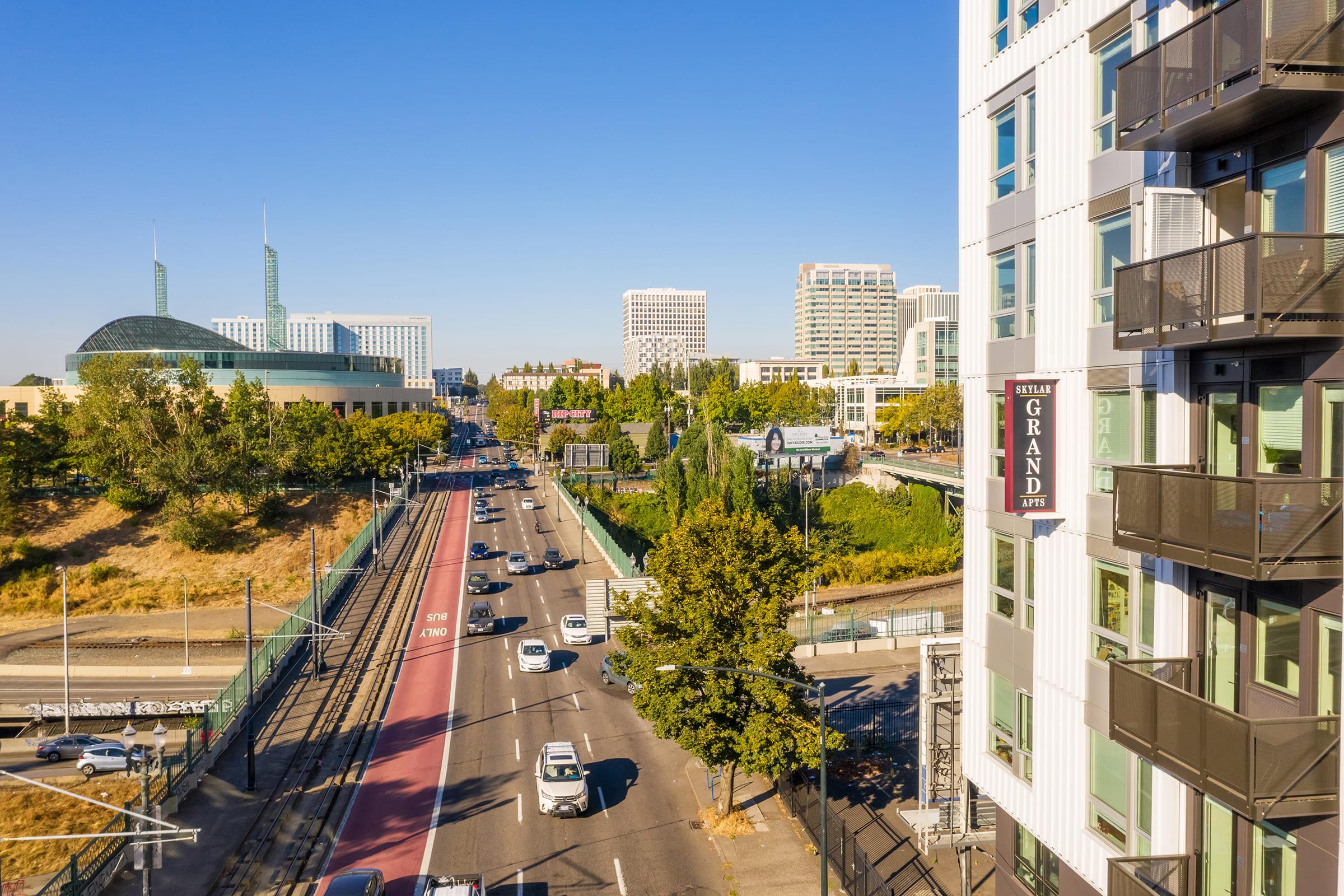 Aerial view of a cityscape featuring a wide street with traffic, lined with trees and modern buildings. Prominent structures include a large, glass-roofed venue on the left and a residential building with balconies on the right. The scene is bathed in bright sunlight against a clear blue sky.