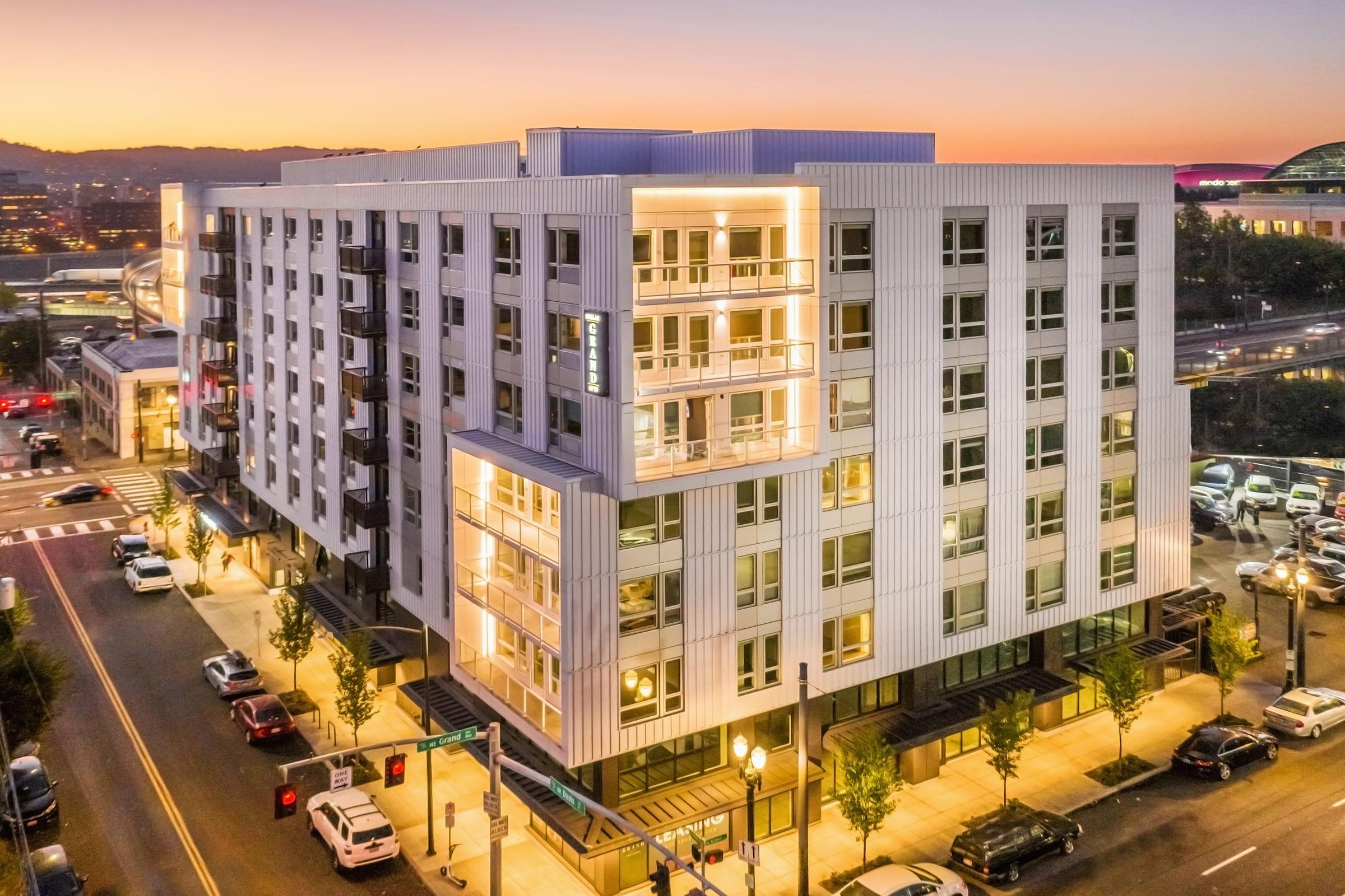 Modern four-story apartment building at sunset, featuring a sleek design with illuminated balconies and large windows. Surrounding streets are lined with trees and parked cars, adding to the urban atmosphere. The sky is a gradient of orange and purple hues.