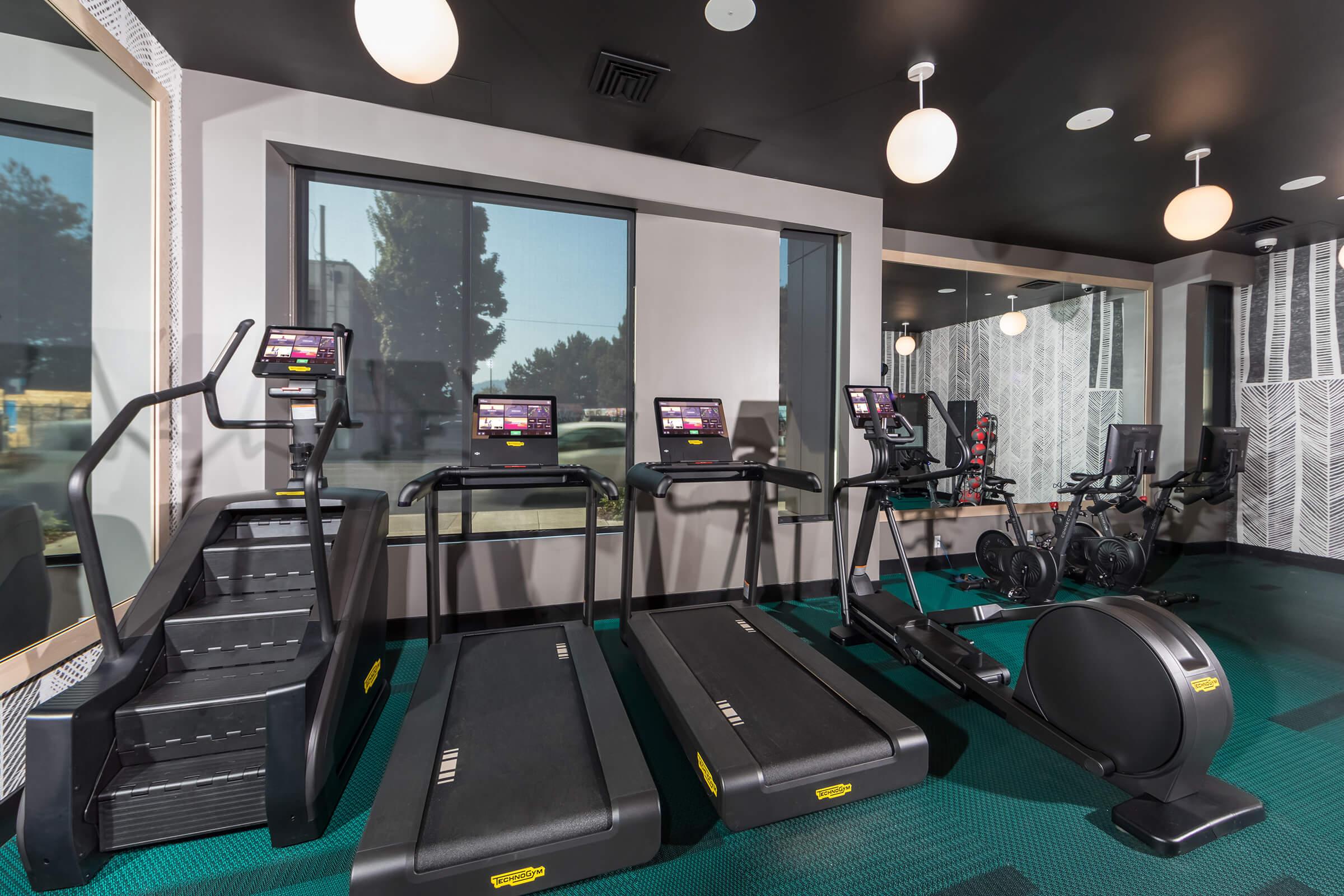 A modern gym interior featuring several fitness machines, including treadmills, an elliptical, and a recumbent bike. The space has large windows allowing natural light, and is designed with sleek, contemporary decor, and bright overhead lighting.