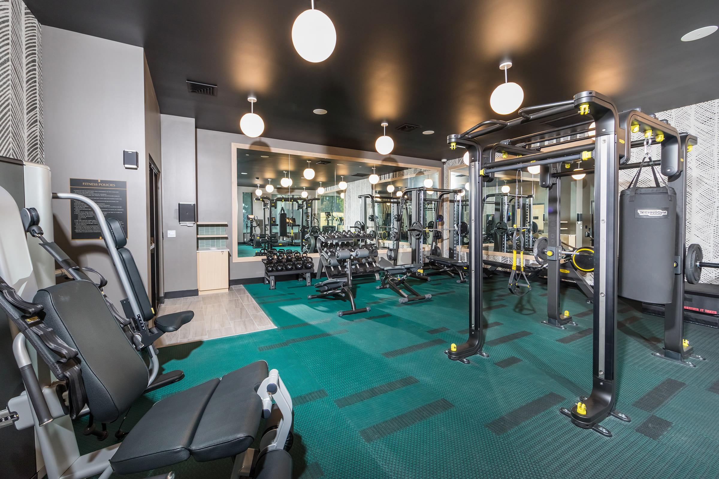 A modern gym interior featuring various exercise equipment, including weight machines and free weights. The floor is covered with teal carpeting, and the space is well-lit with circular pendant lights. Mirrors line one wall, reflecting the equipment and enhancing the overall brightness of the room.