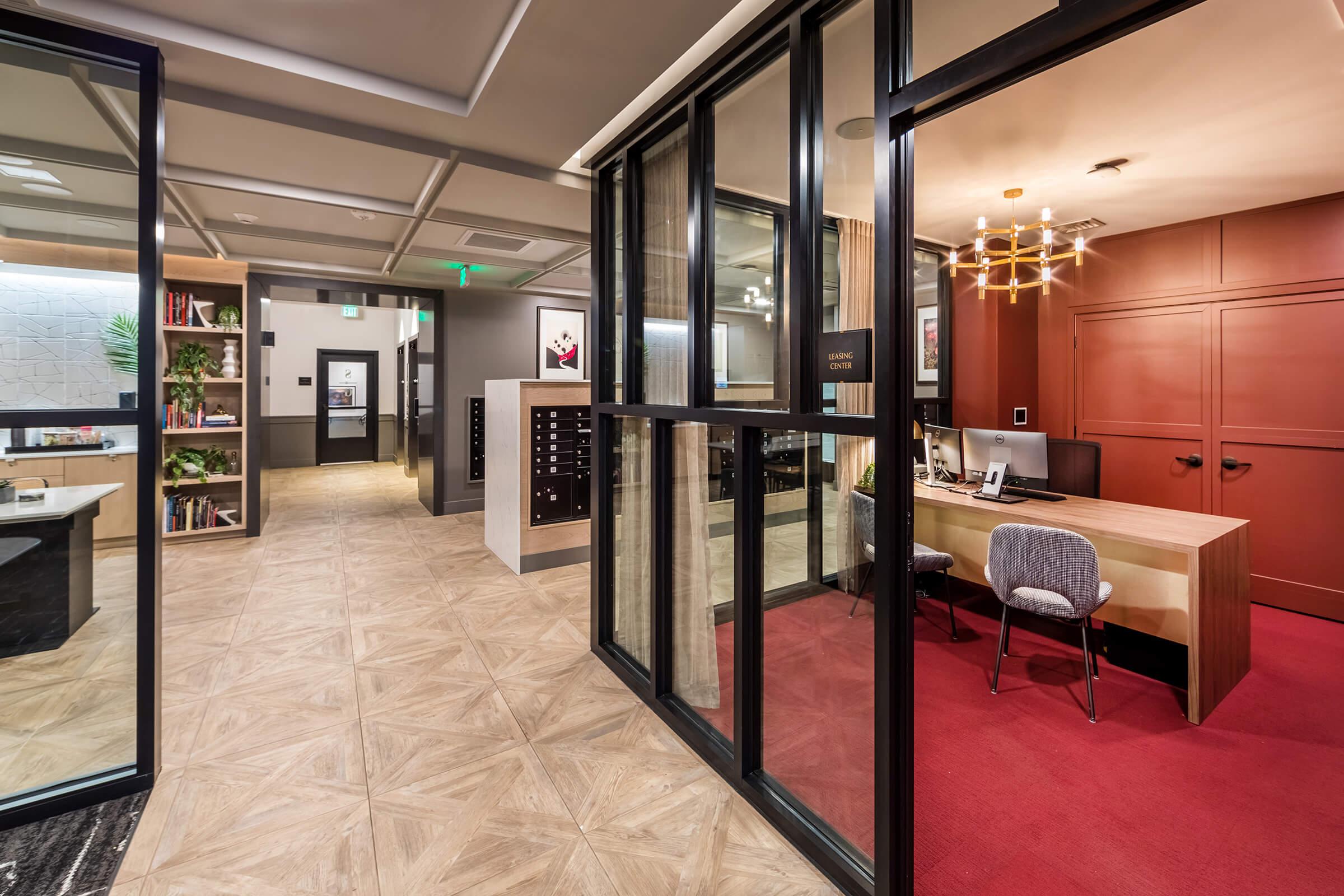 Interior view of a modern office space featuring a reception area with a wooden desk and a chair. The room has large glass doors, contemporary lighting, and decorative elements. A corridor leads to additional rooms, with a mix of textures and colors, including red carpeting and wooden flooring.
