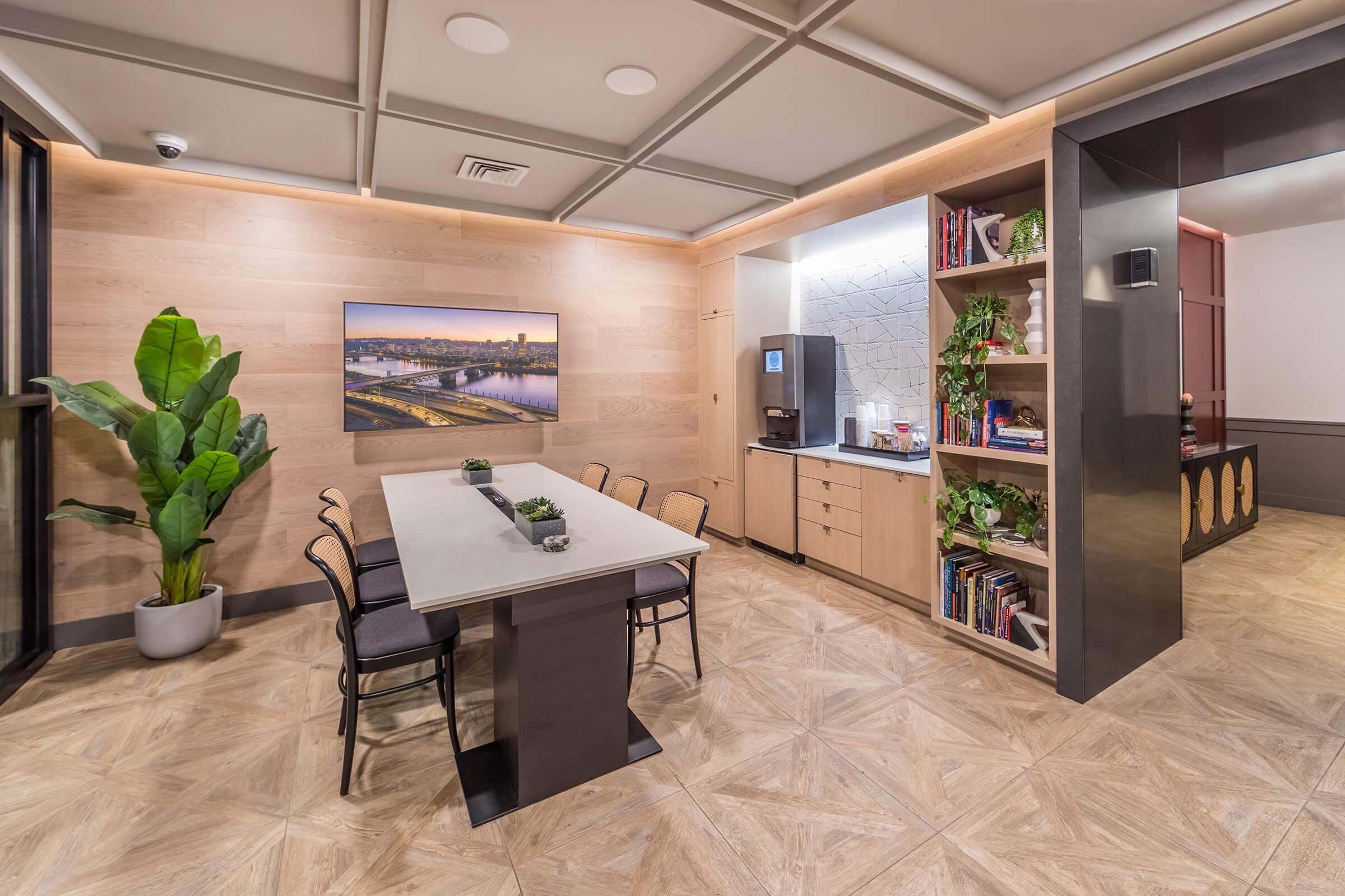 Modern kitchen and dining area featuring a rectangular table with black chairs, surrounded by warm wood walls. A tall plant adds greenery, while a kitchenette with a coffee machine and shelves filled with books and plants can be seen in the background. Soft lighting enhances the inviting atmosphere.