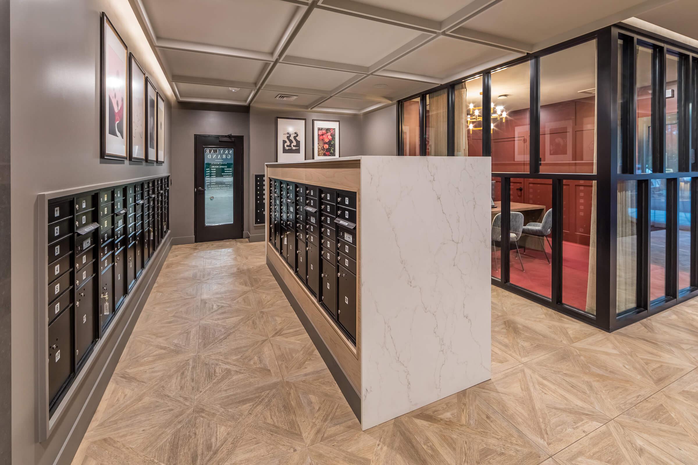 A modern mailroom with a marble partition featuring multiple mailboxes on one side. The walls are decorated with framed artwork, and a glass-walled room is visible in the background. The floor has a patterned design, and a door leads to the outside.