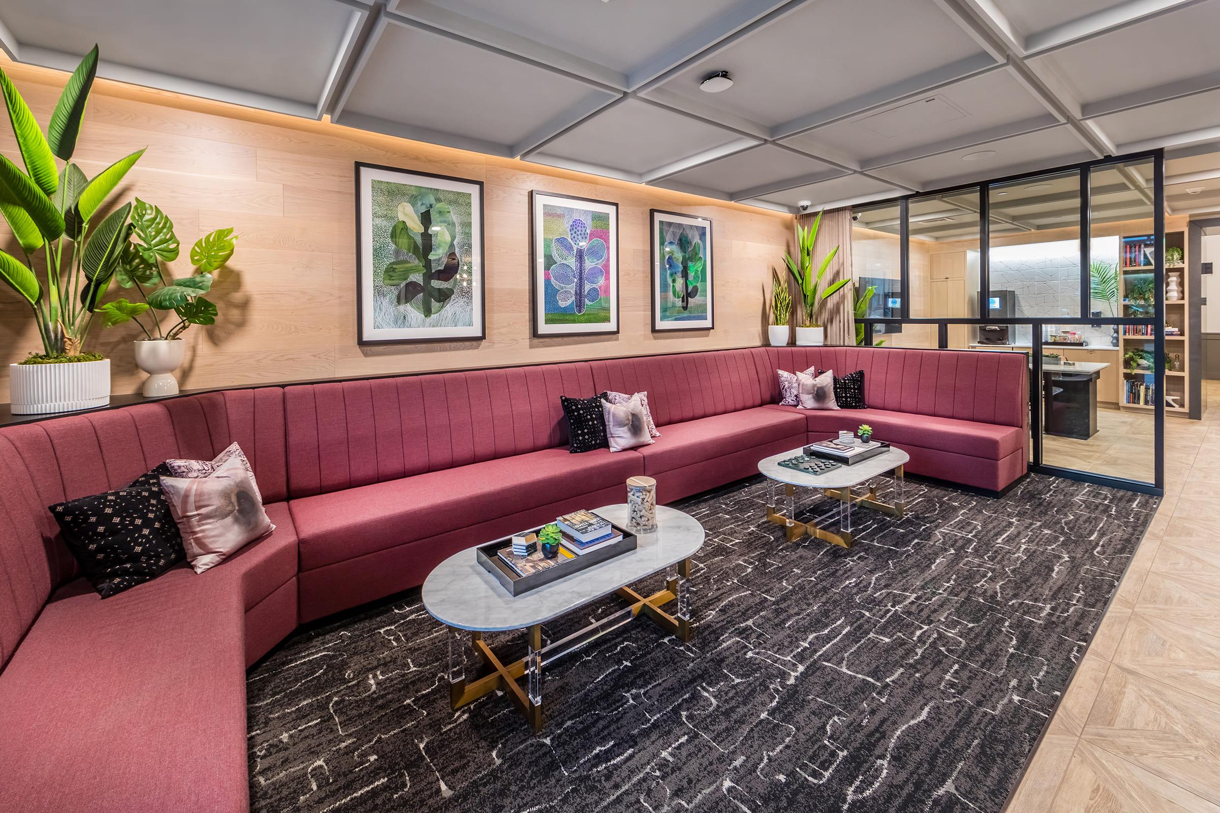 A modern lounge area featuring a large pink sectional sofa with decorative pillows, two coffee tables with books and decor items, and plants. The walls are adorned with framed artwork. A glass partition reveals a workspace in the background. The floor has patterned tiles, adding to the contemporary aesthetic.