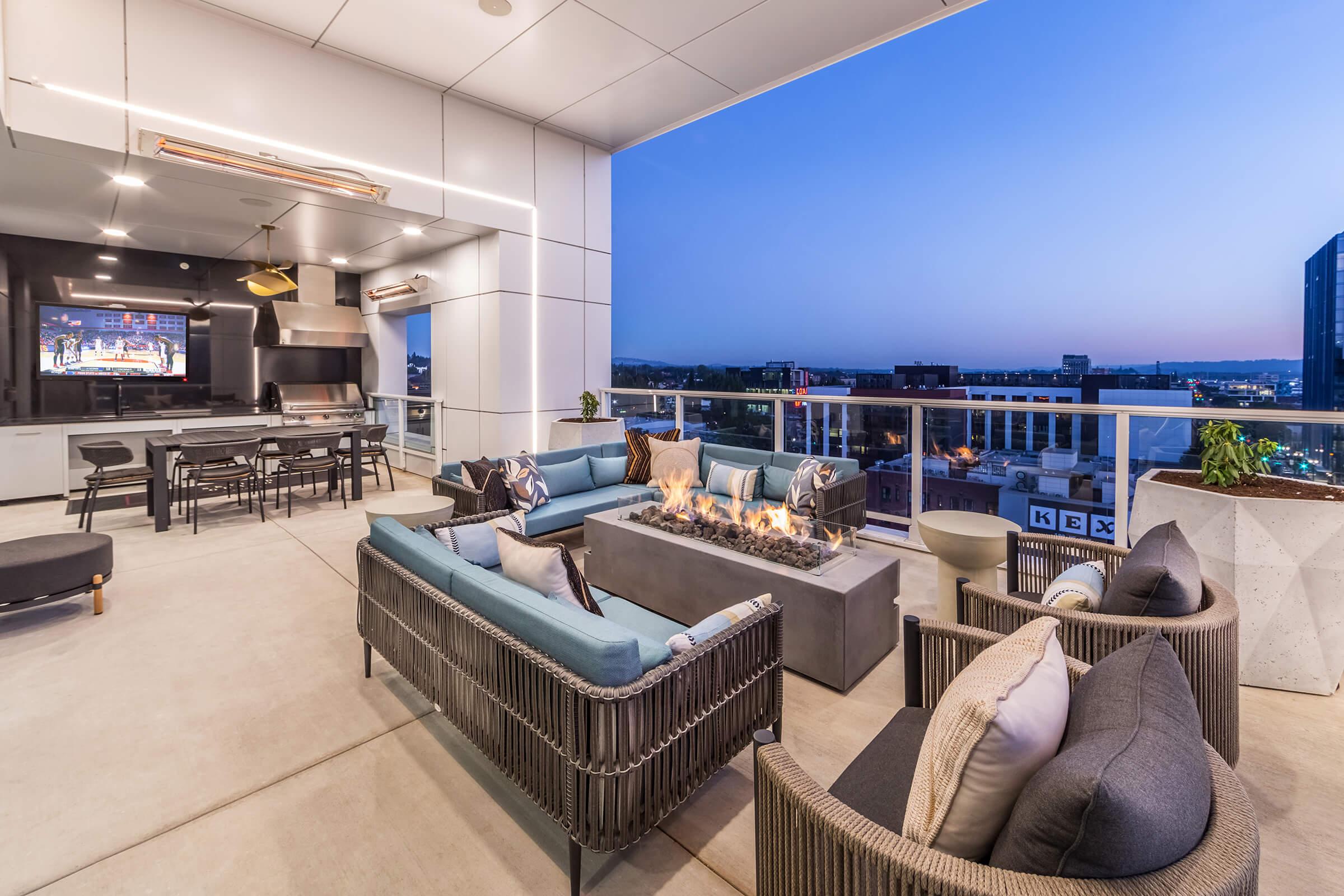 A stylish outdoor patio with modern furniture, including a plush seating area surrounding a fire pit. An outdoor dining set is visible, along with a wall-mounted TV. The setting is illuminated by ambient lighting, with a beautiful twilight skyline in the background.