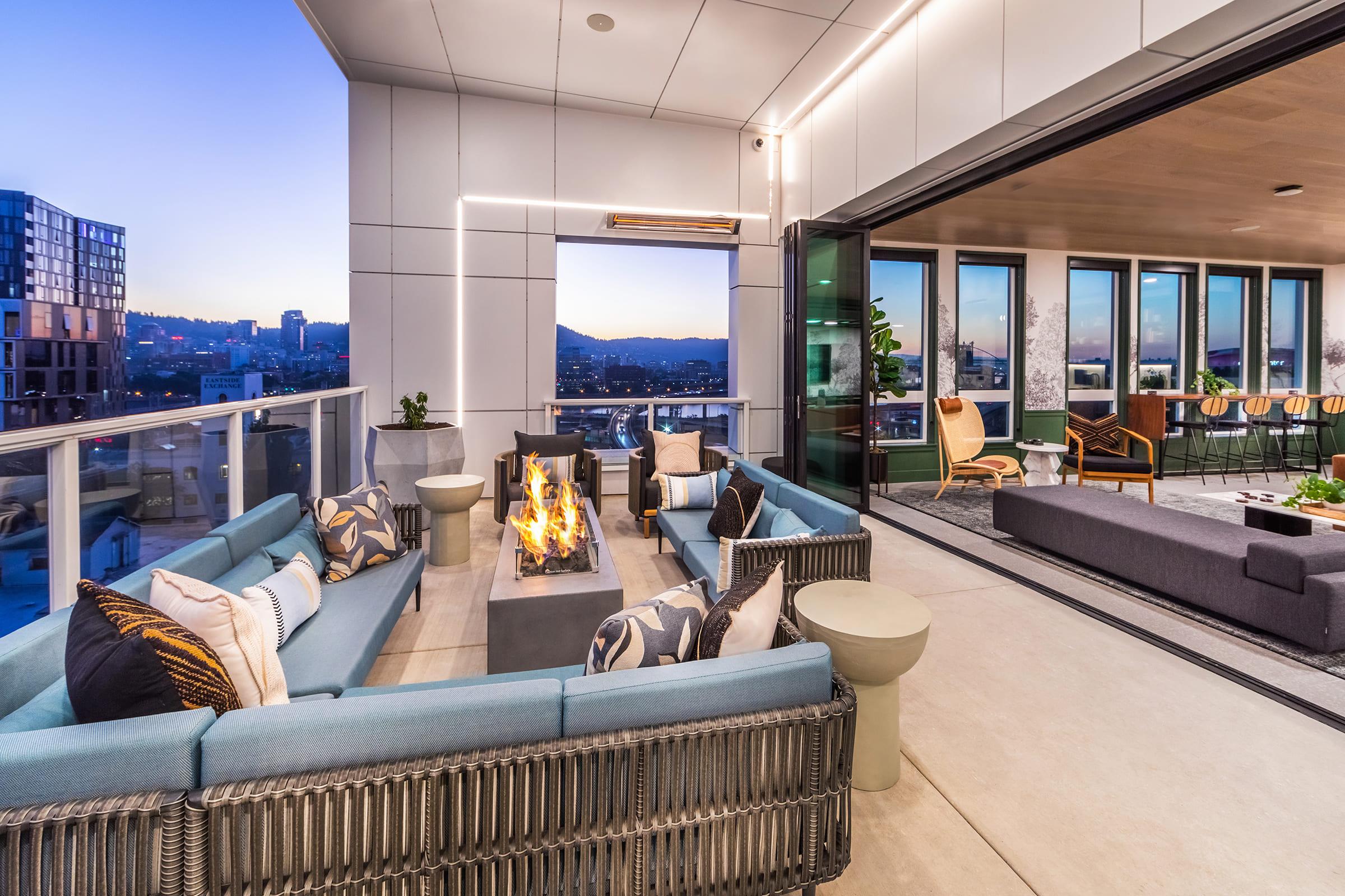A modern rooftop terrace featuring comfortable seating arrangements with blue cushions, a central fire pit with flames, and glass railings. The background showcases a city skyline at sunset, with large windows opening to a stylish interior space adorned with greenery.
