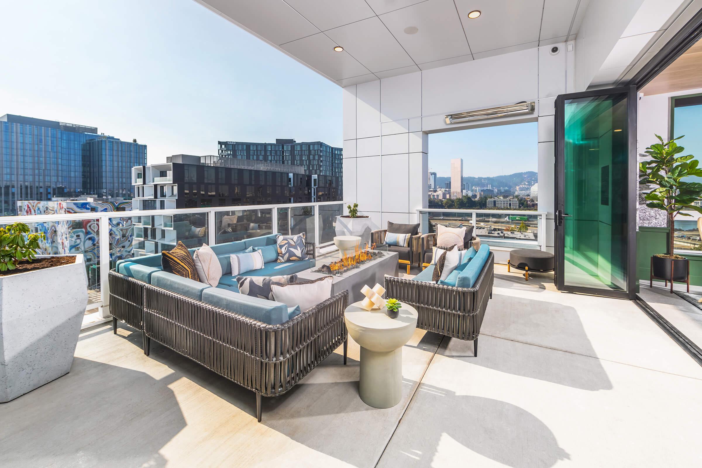 Modern outdoor terrace featuring a stylish seating area with plush cushions, a coffee table, and potted plants. The space offers a panoramic view of the city skyline and surrounding buildings, with glass sliding doors leading to an interior area. Bright sunlight illuminates the scene.