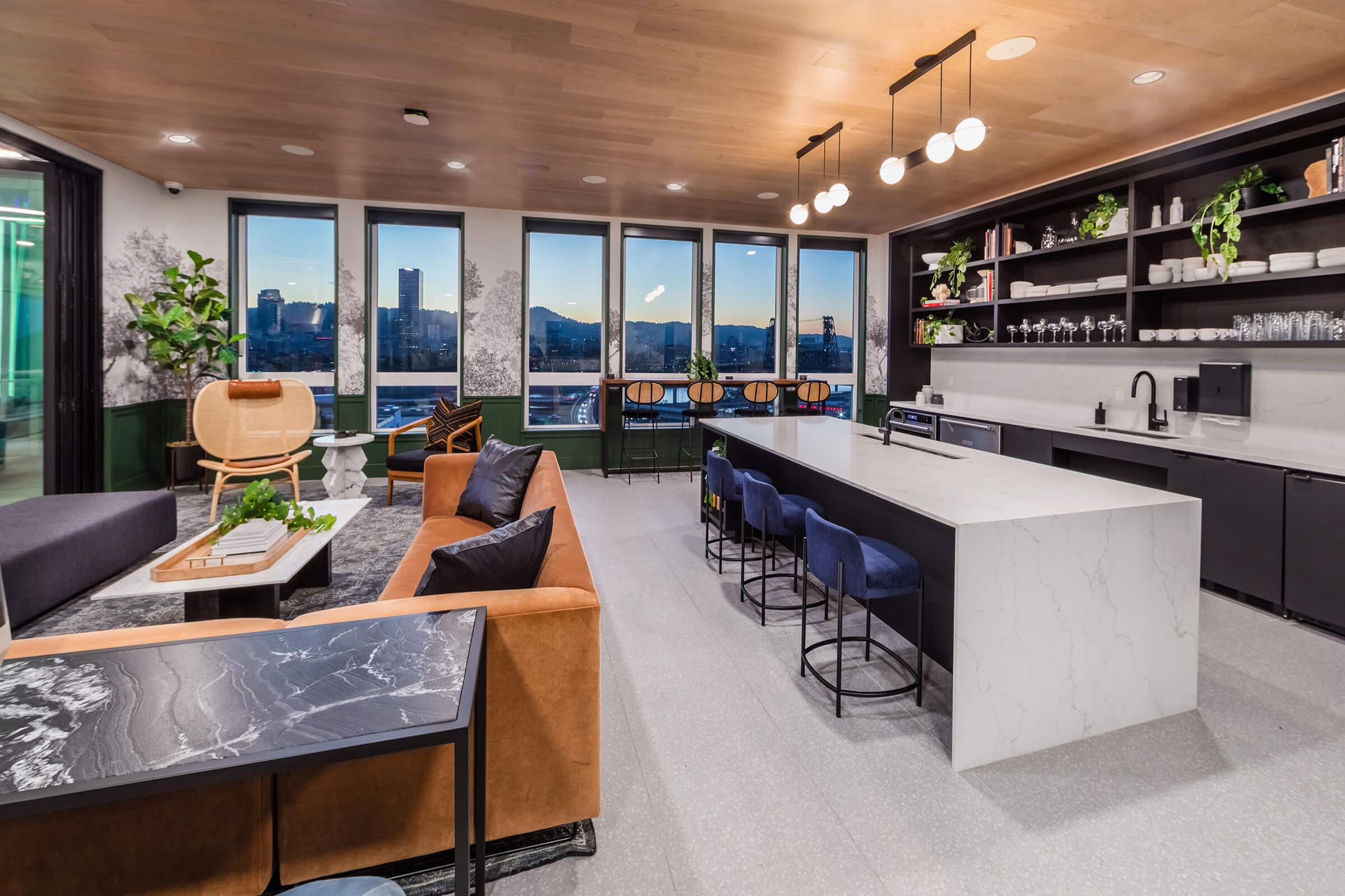 Modern apartment lounge with large windows offering city views. Features a mix of seating, including a brown sofa, wooden chairs, and bar stools. A sleek white island counter with black accents contrasts a dark, textured table. Potted plants add greenery to the stylish, contemporary decor.
