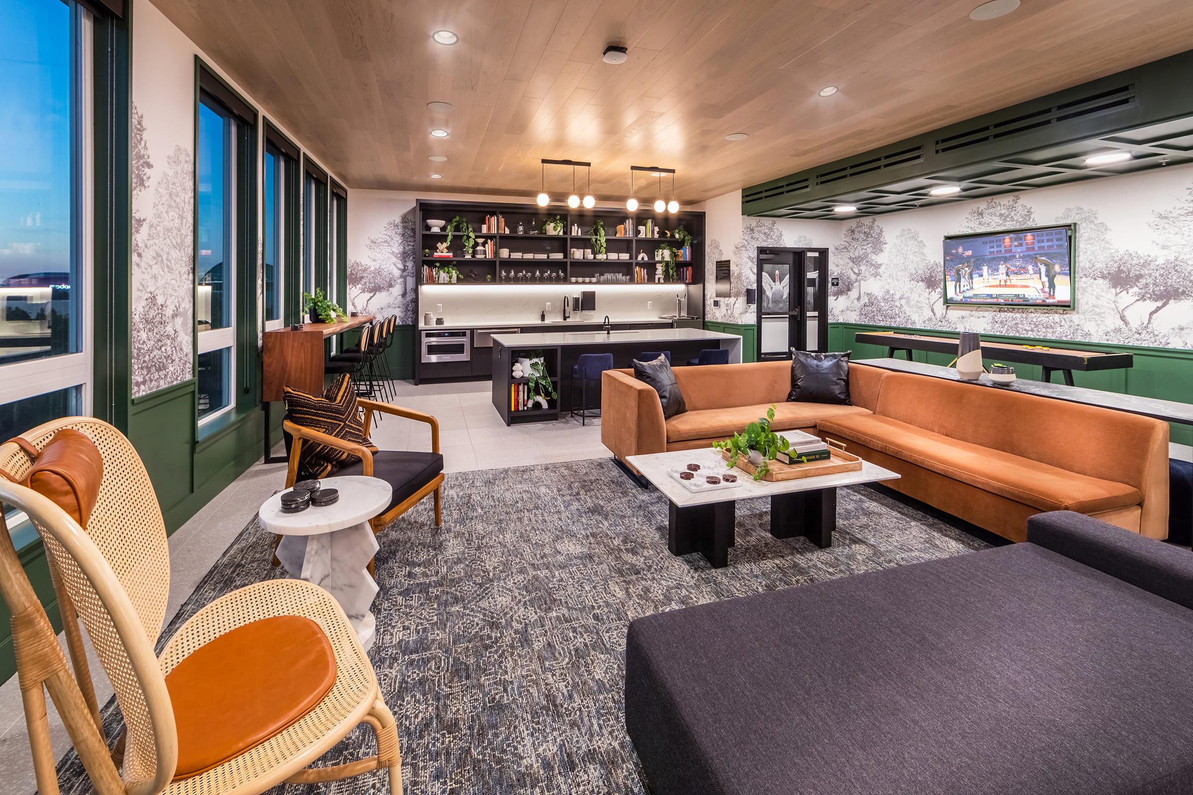 A modern lounge area featuring a mix of seating, including a brown sofa, black couch, and rattan chairs. The space has large windows allowing natural light, a stylish kitchen area with dark cabinetry, and decorative plants. A large TV is mounted on the wall, completing the inviting atmosphere