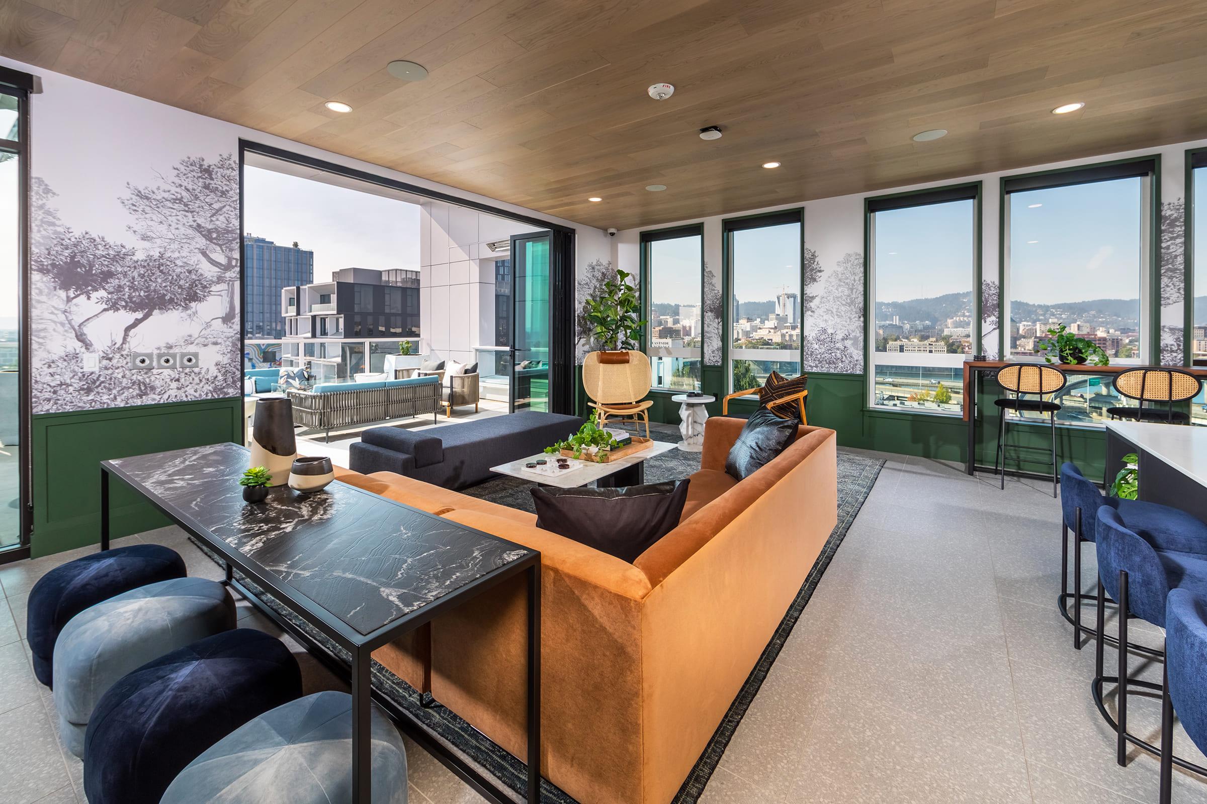 A modern, stylish living area featuring a large brown sofa, a sleek black dining table with stools, and large windows that provide a view of the city skyline. The space is decorated with plants and contemporary furnishings, creating a bright and inviting atmosphere.