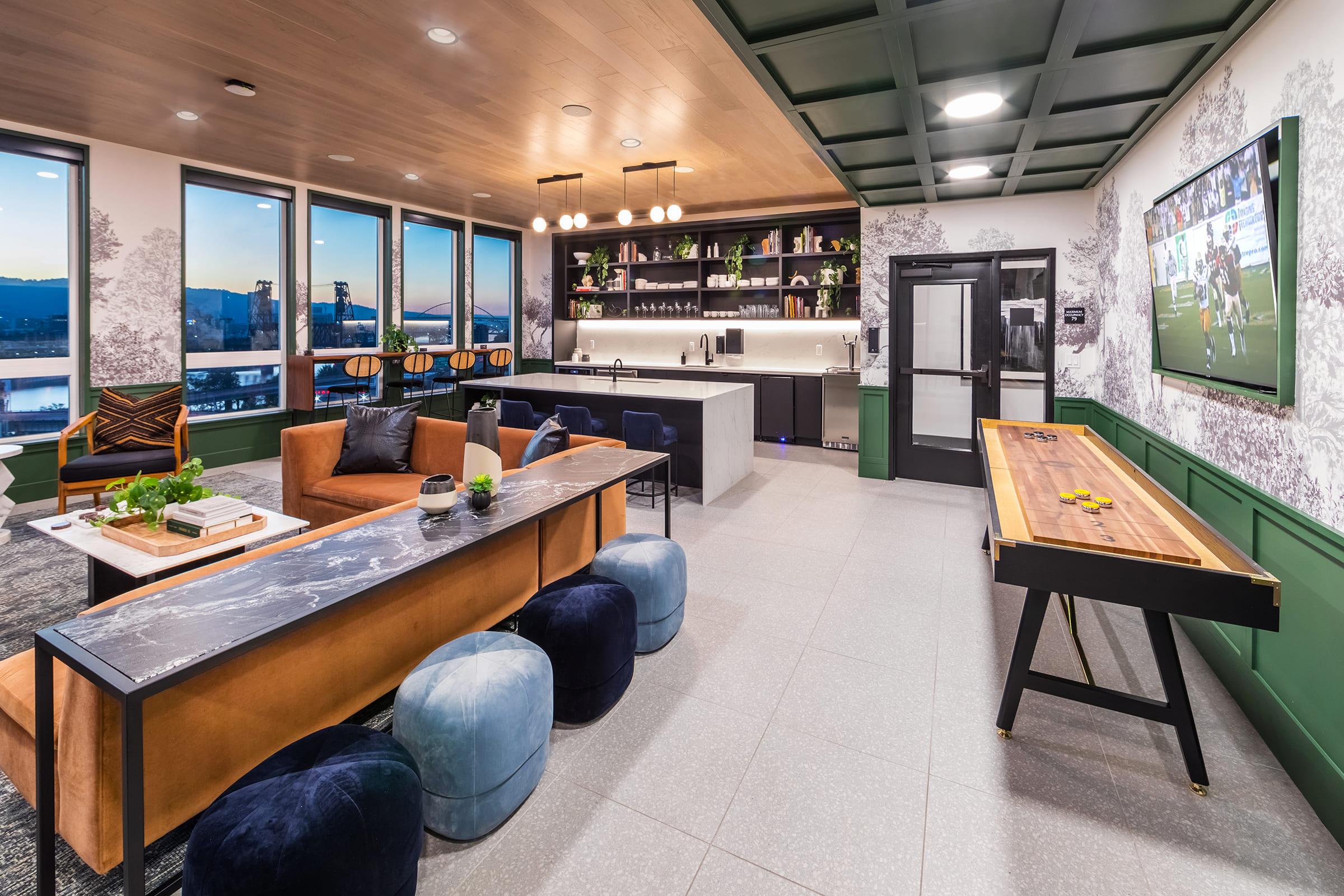 A modern communal lounge featuring a sleek kitchen with bar seating, a shuffleboard table, and comfortable seating areas. Large windows showcase a panoramic view, while stylish decor includes patterned wallpaper and greenery. A screen displays a sports game in the background, enhancing the inviting atmosphere.