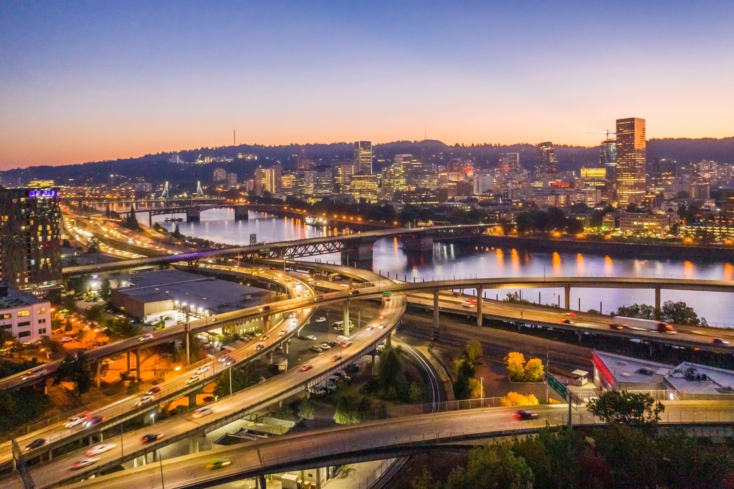 Aerial view of a cityscape at dusk, showcasing a network of highways and bridges over a river, with the skyline of Portland illuminated in the background. The scene captures a mix of urban structures, soft sunset colors, and traffic movement, creating a vibrant evening atmosphere.