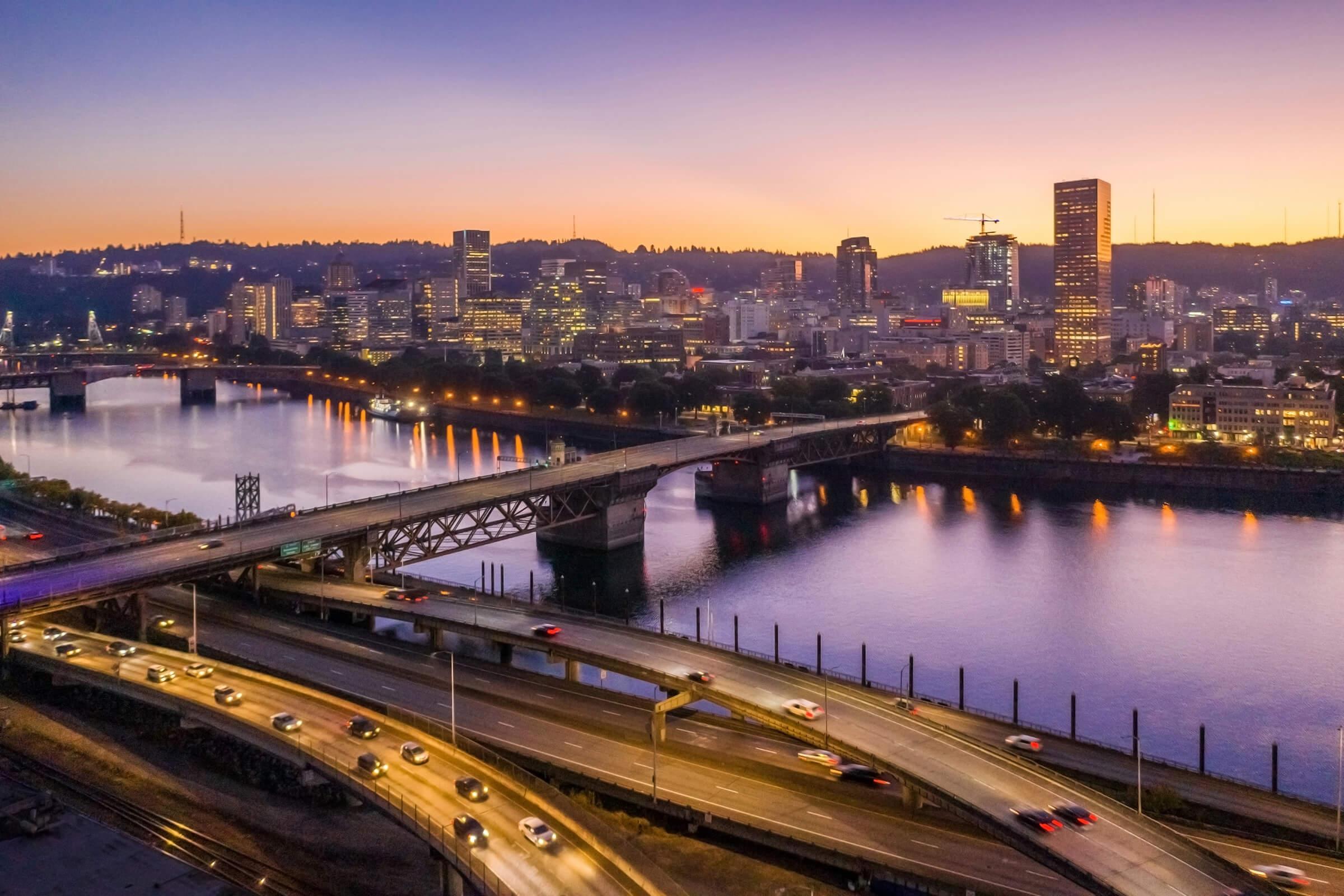 A panoramic view of a city skyline at sunset, featuring a river and bridge in the foreground. The sky is a gradient of purple and orange hues, with city buildings illuminated. Traffic can be seen on the roads, and the water reflects the colorful sky and city lights.