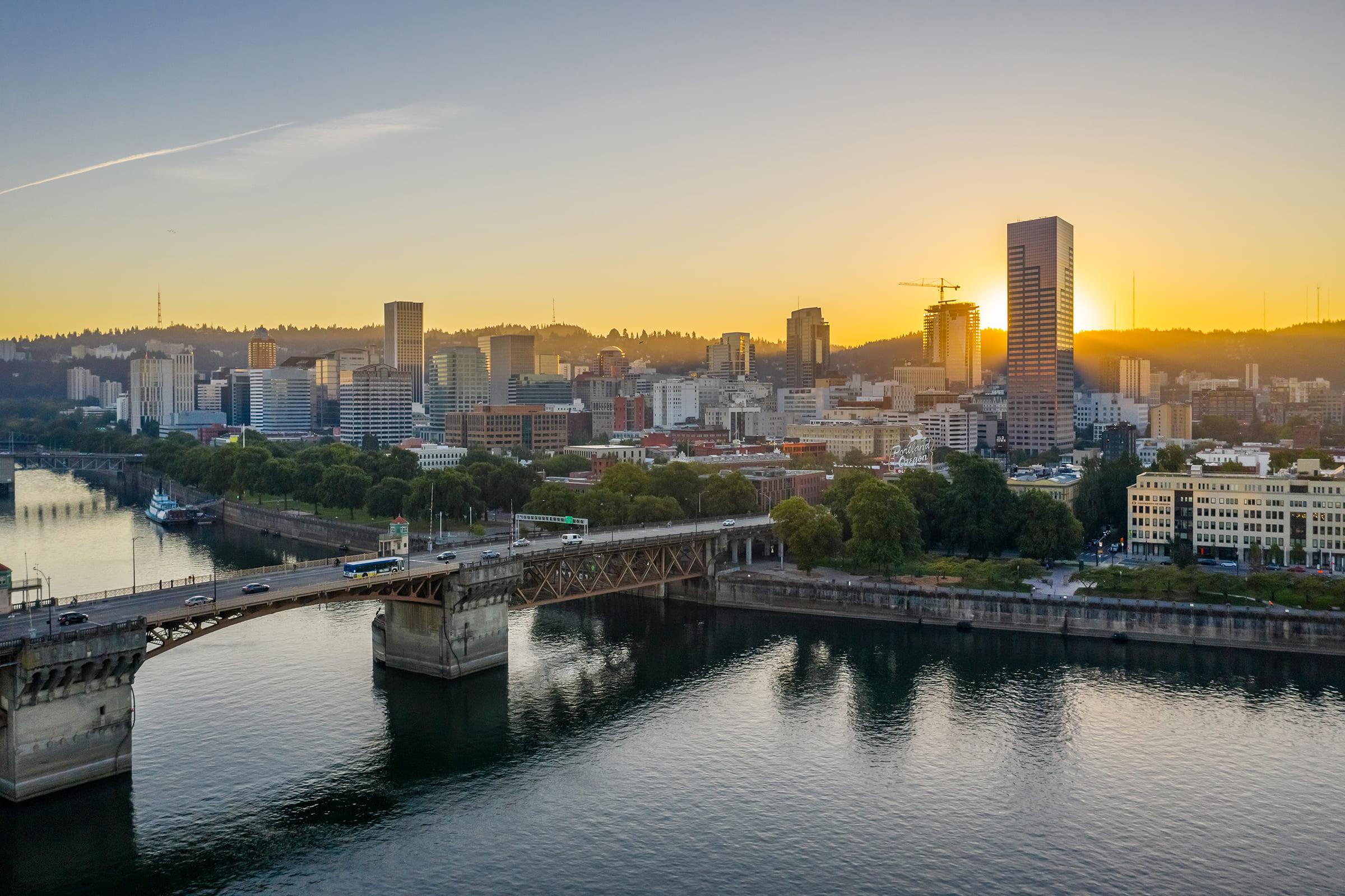 A scenic view of a city skyline at sunset, featuring tall buildings and a bridge over a river. The sun casts a warm glow, illuminating the skyline and creating reflections on the water. Trees line the riverbank, adding greenery to the urban landscape.