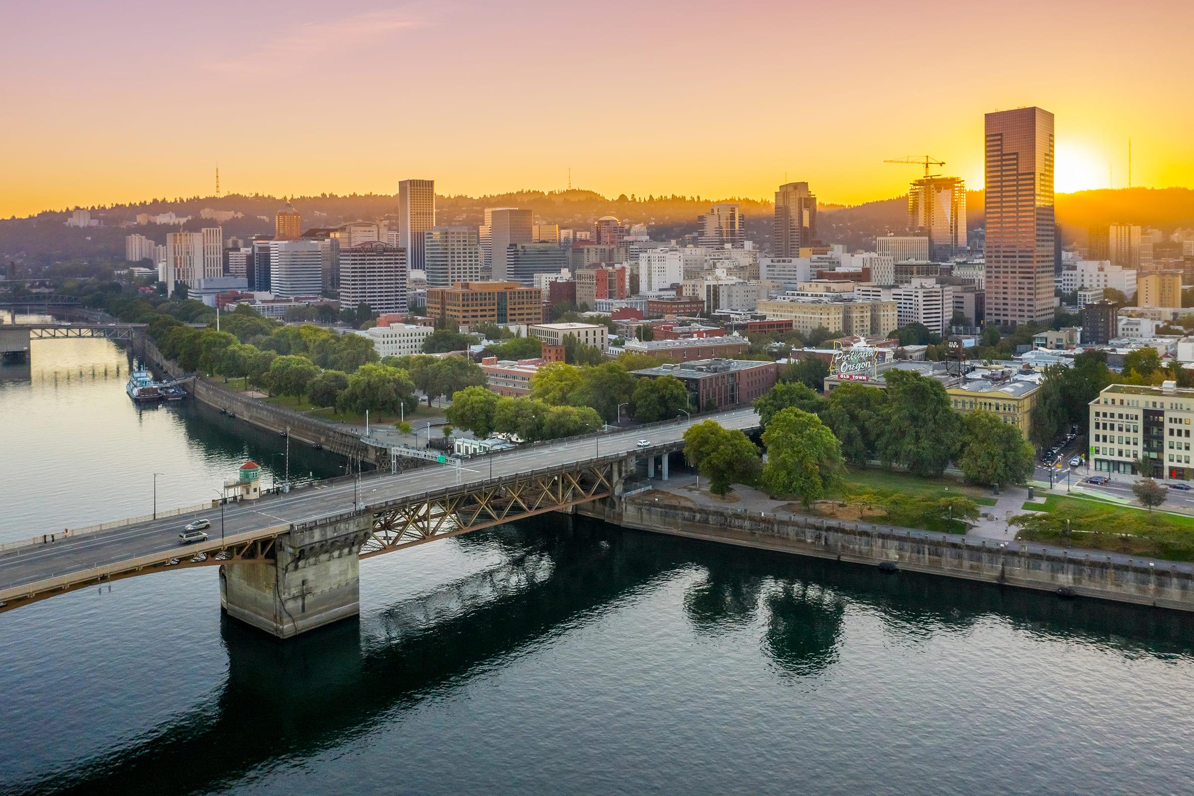 Aerial view of a city's skyline at sunset, featuring tall buildings and a river reflecting the warm colors of the sky. A bridge spans the river, while trees line the banks. Construction cranes are visible, indicating ongoing development. The scene conveys a sense of urban life and natural beauty.