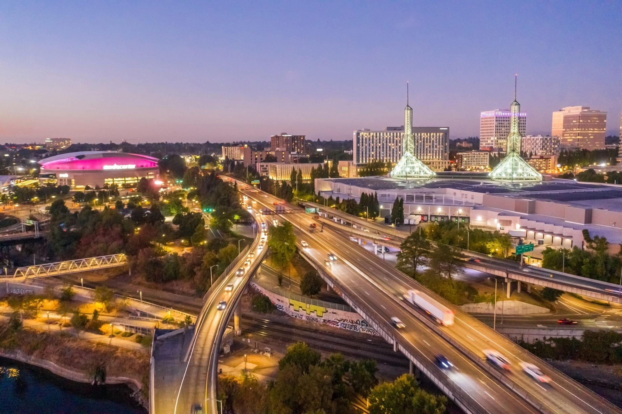 Aerial view of a city at dusk, showcasing a busy highway with vehicles, modern buildings, and illuminated structures. The skyline features unique architectural towers and a vibrant entertainment venue with bright pink lighting, surrounded by greenery and urban infrastructure.