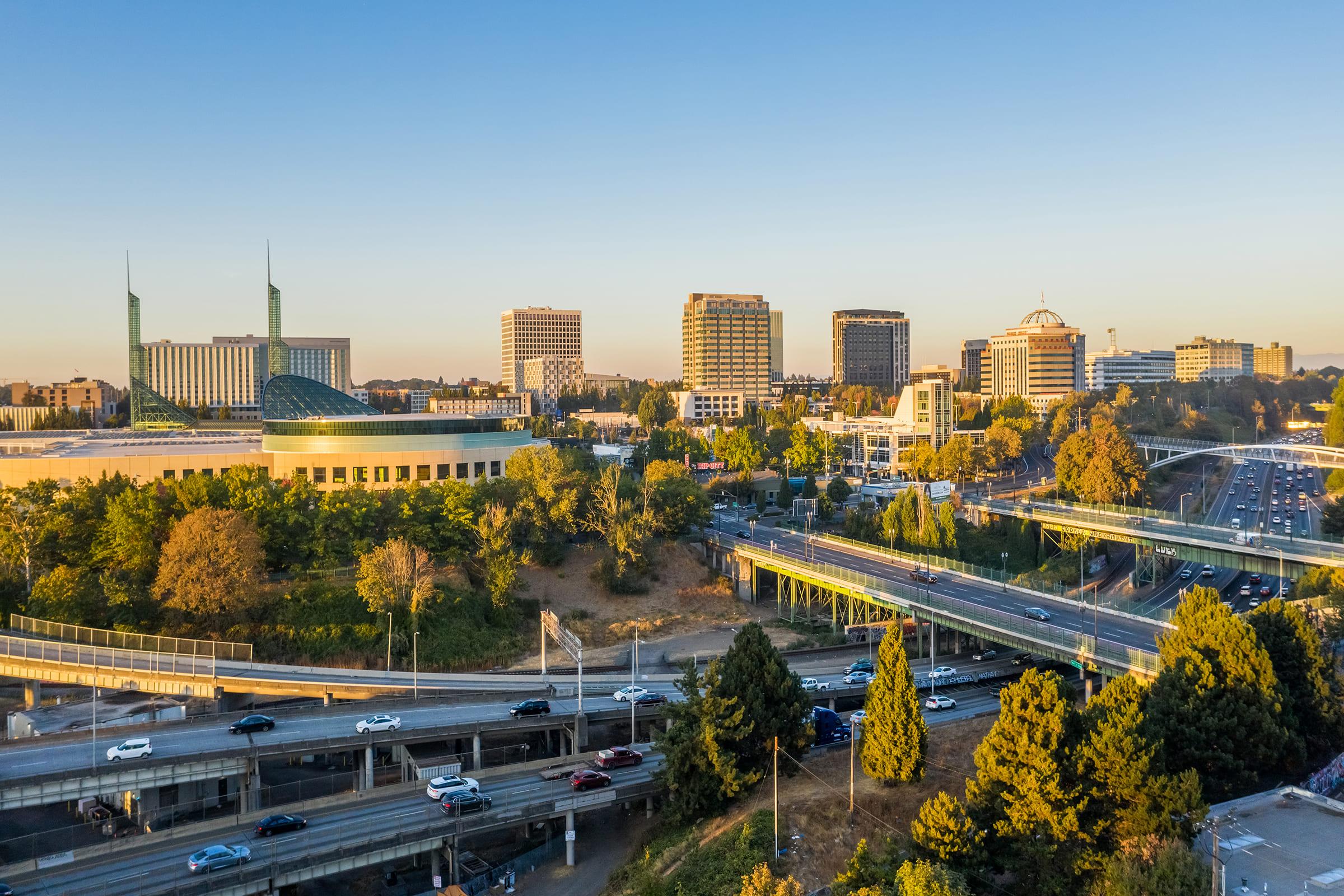 A scenic view of a downtown urban area at sunset, featuring modern high-rise buildings, a large dome structure, and major roadways with vehicles. Lush trees and greenery are visible in the foreground, providing a contrast to the cityscape. The sky is clear, showcasing warm golden hues.