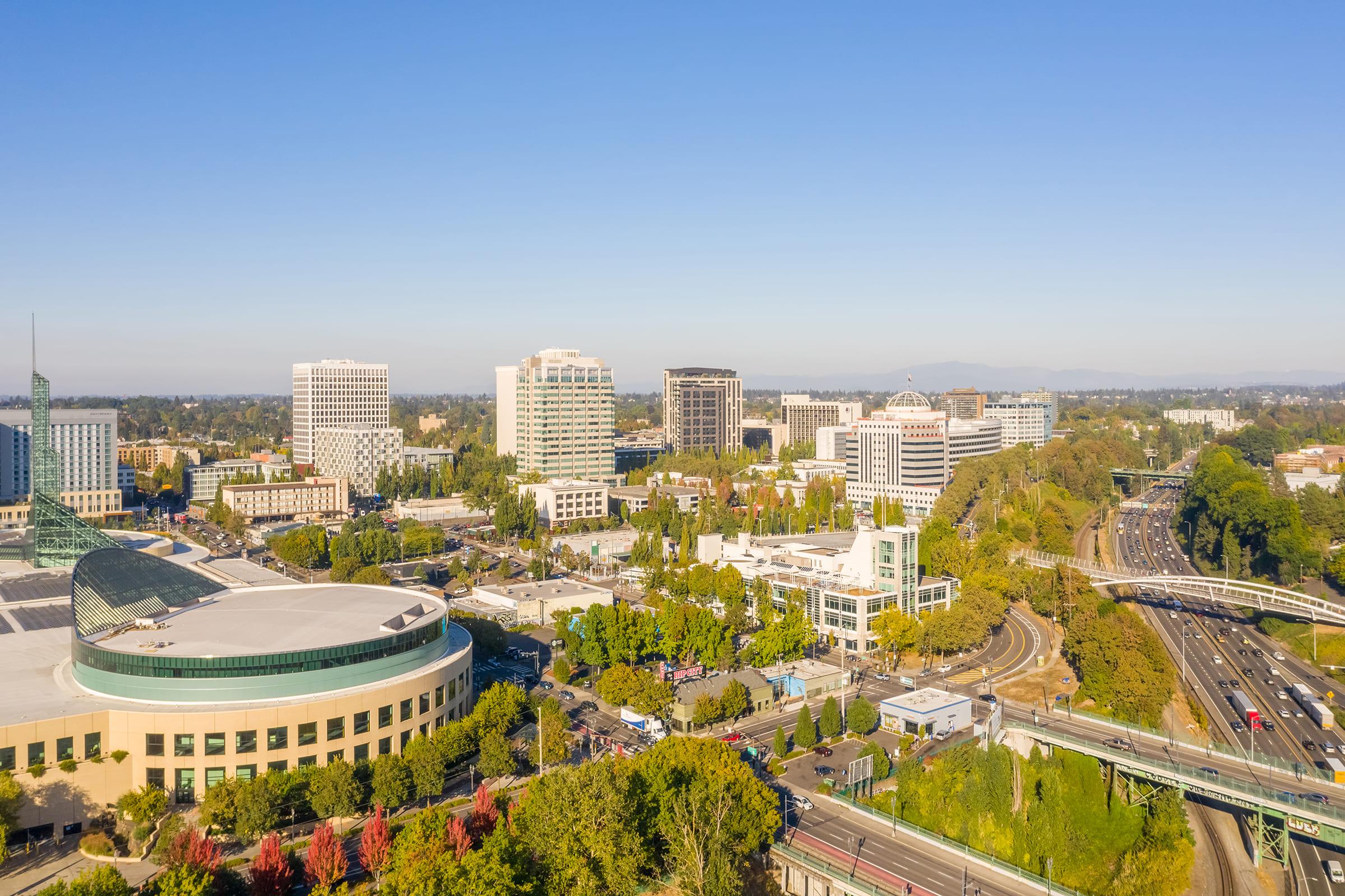Aerial view of a modern urban landscape featuring high-rise buildings, a convention center, and a highway. The scene is set under a clear blue sky, surrounded by greenery and a mix of commercial and residential structures, indicating a vibrant city environment.
