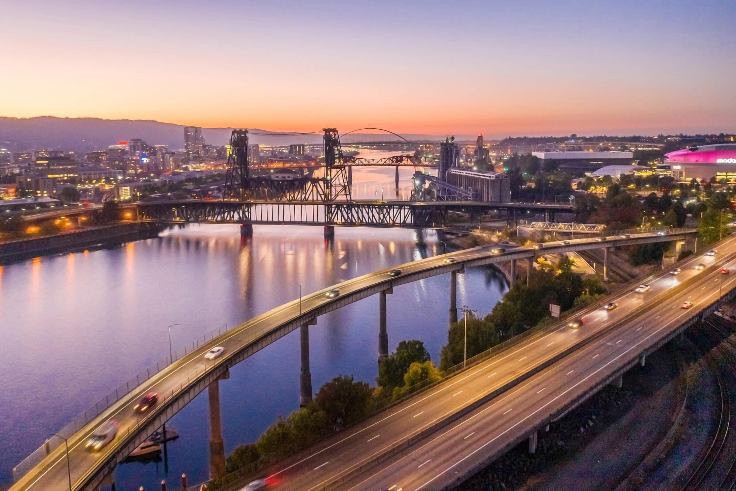 Aerial view of a river with bridges at dusk, showcasing city lights reflecting on the water. Cars travel on nearby highways, and silhouettes of buildings and structures are visible against a gradient sky transitioning from orange to purple.