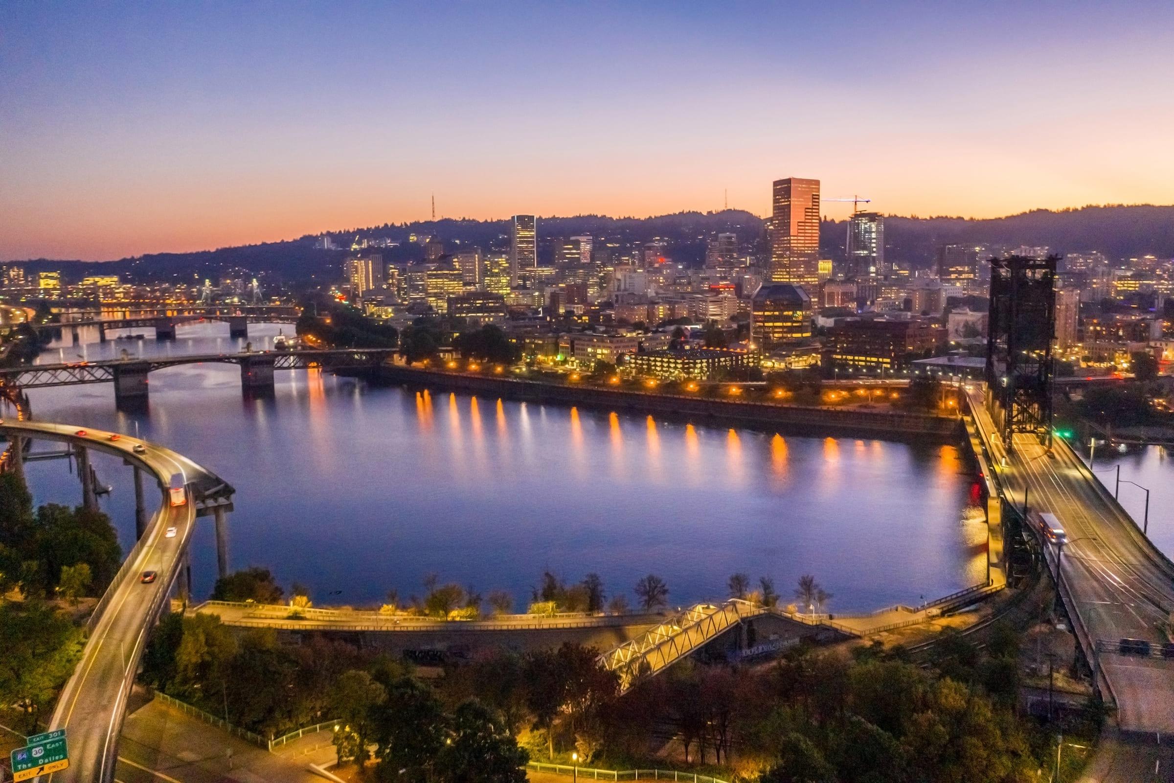 A stunning aerial view of a city skyline at dusk, showcasing illuminated buildings reflecting on a calm river. The scene features bridges crossing the water, with a winding road in the foreground and hills in the background, creating a serene urban landscape.