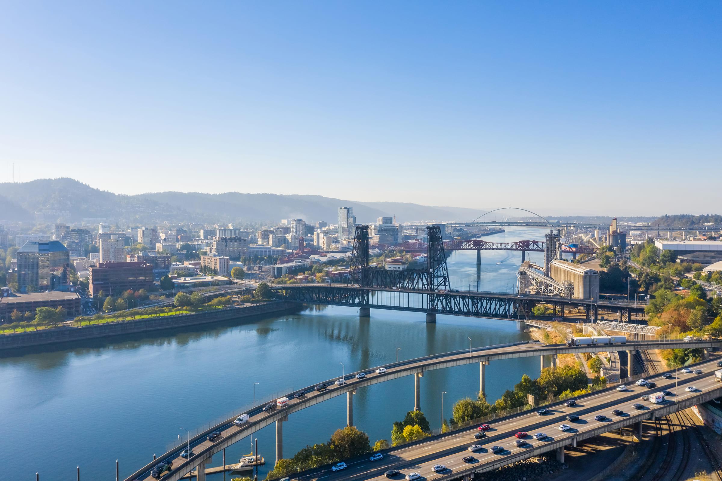 Aerial view of a cityscape featuring a river with bridges, surrounded by hills and buildings. Clear blue sky above, showcasing urban infrastructure along the waterfront with vehicles on a highway. The scene captures a vibrant, sunny day in an urban area, likely a city near a river.