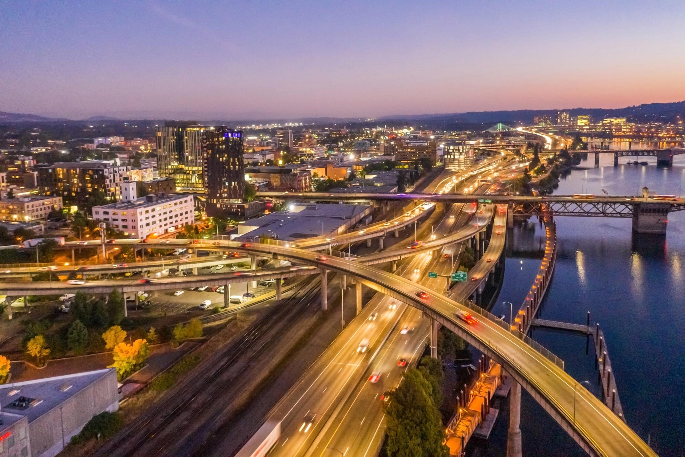 Aerial view of a cityscape at dusk, featuring busy highways with flowing traffic, a river, and illuminated buildings. The sky transitions from orange to deep blue as night approaches, showcasing the vibrant urban environment.