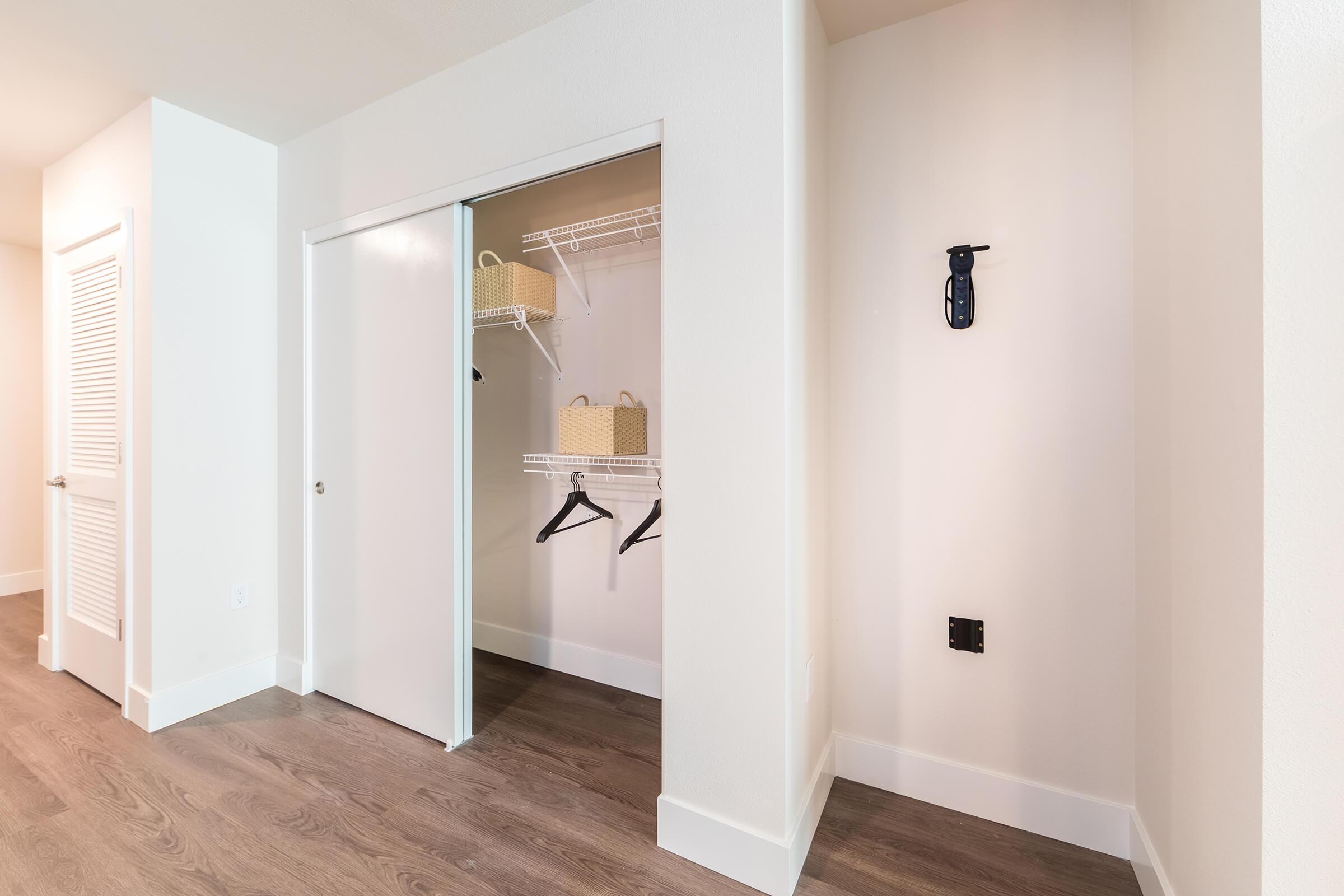 Interior view of a modern room featuring a sliding closet door, showcasing organized shelving with hanging space and storage baskets. Light-colored walls and wooden flooring enhance the bright, spacious feel. A small wall mount is present beside the closet.