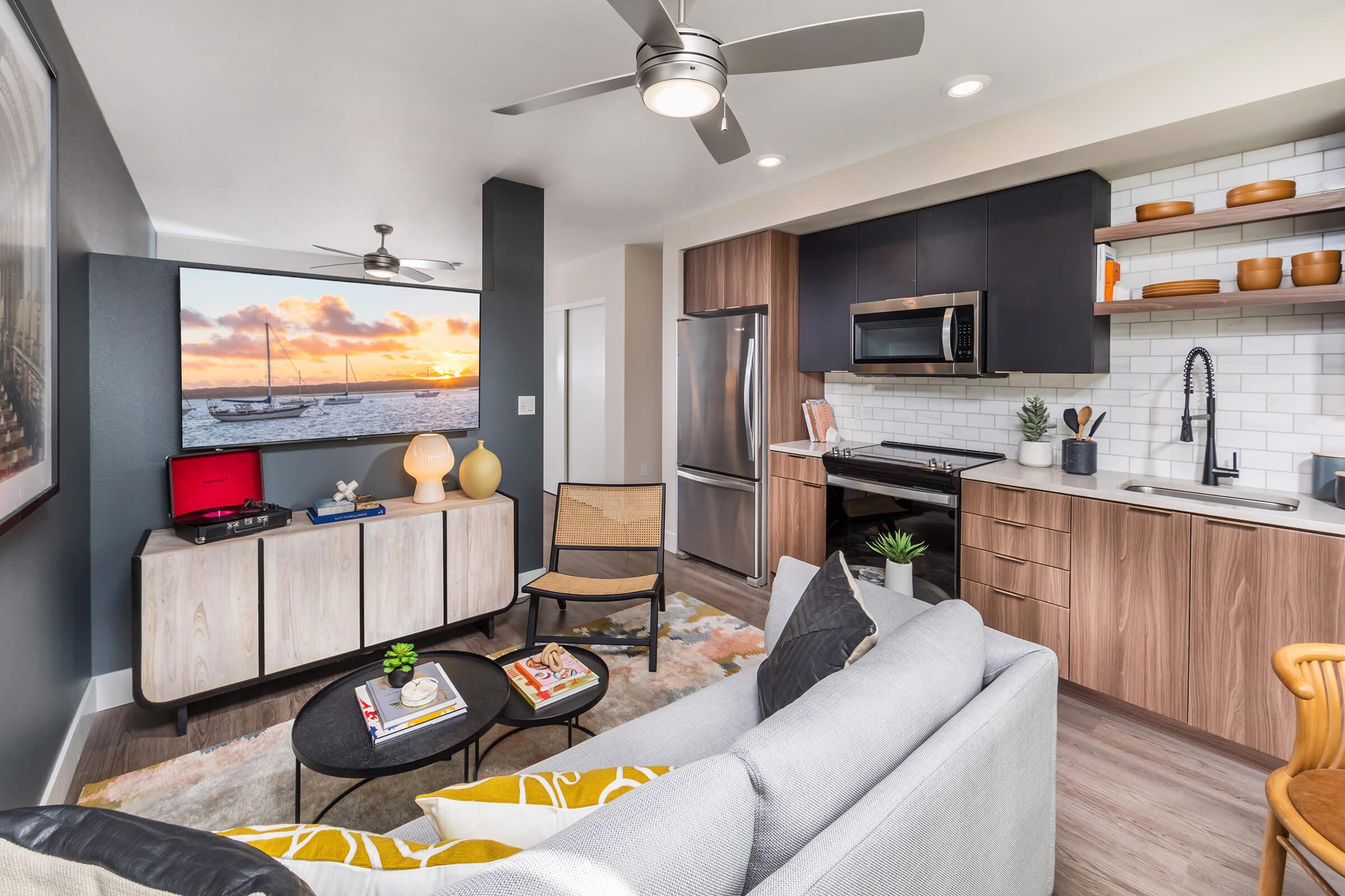 A modern living space featuring a cozy gray sofa with yellow accent pillows, a black coffee table, and a stylish wood cabinet. The kitchen has stainless steel appliances and stylish decor. A wall-mounted television displays a sunset over water, enhancing the inviting atmosphere.