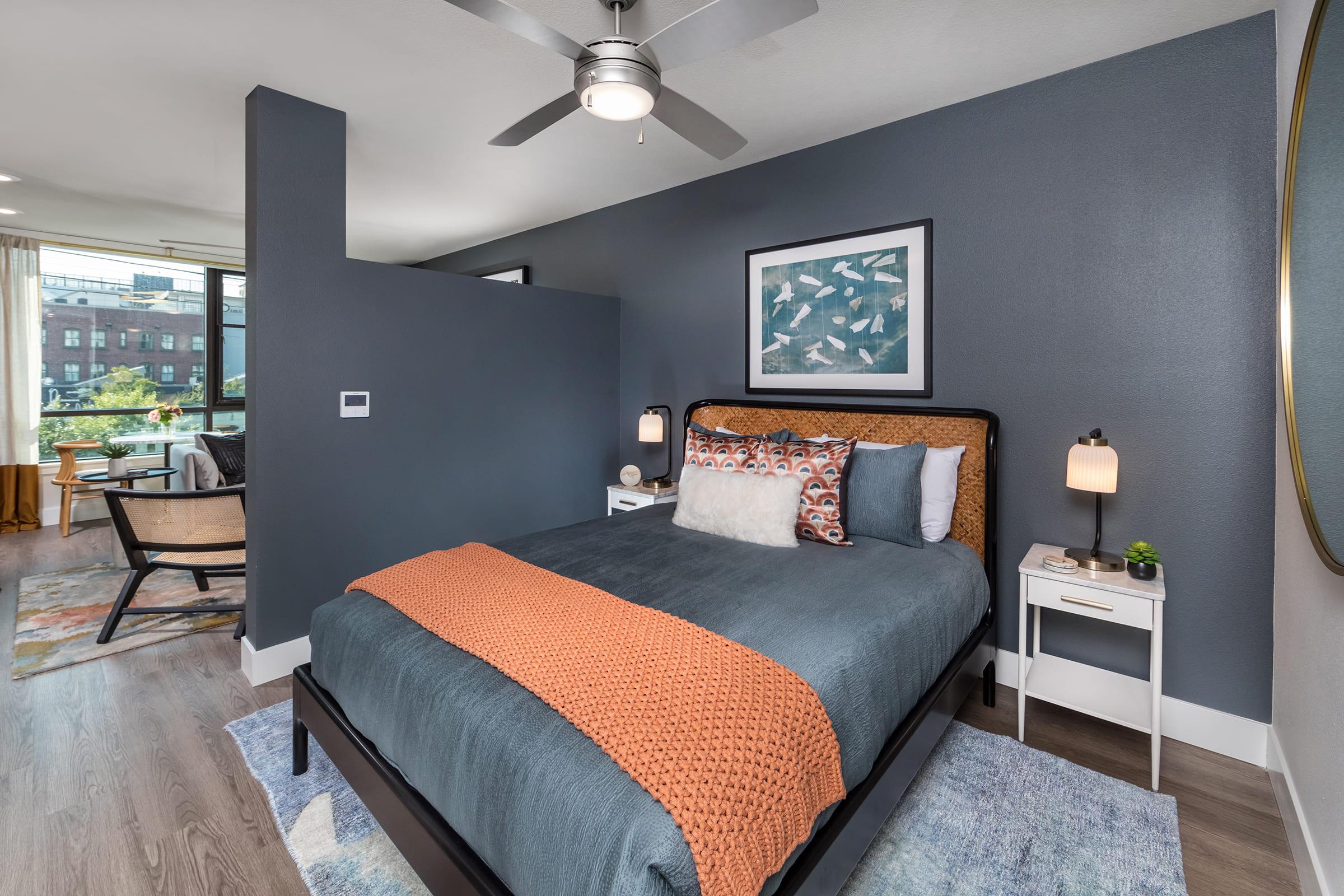 A modern bedroom featuring a comfortable bed with an orange throw, decorative pillows, and stylish bedside lamps. The dark blue accent wall adds a contemporary touch. A round mirror hangs above a nightstand, while a cozy chair and window reveal a view outside, enhancing the inviting atmosphere.