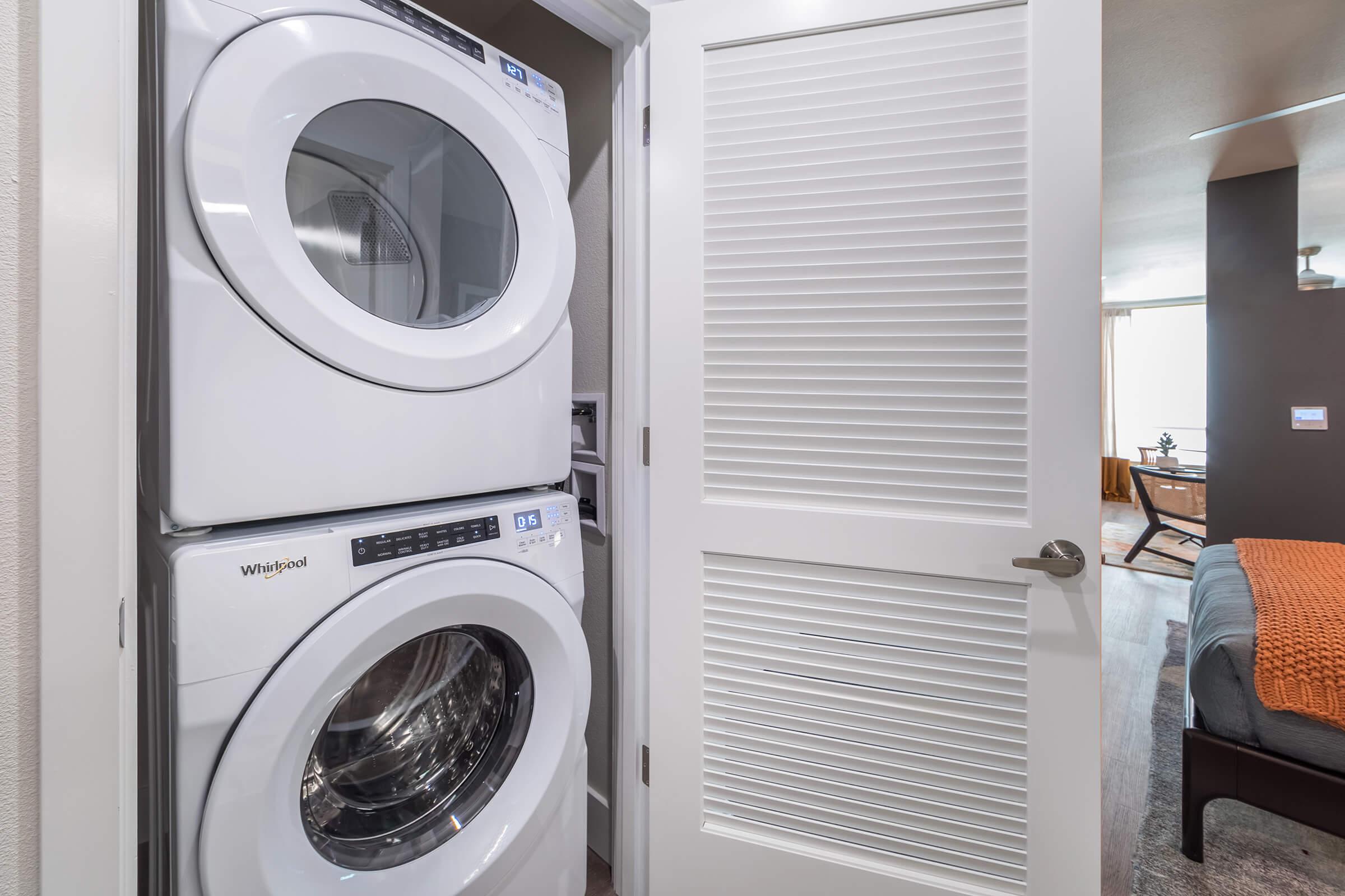 A stacked washer and dryer from Whirlpool located in a laundry nook, partially visible through a closed door. The surrounding area features a modern interior with a warm-toned blanket on a nearby couch and natural light coming from a window.