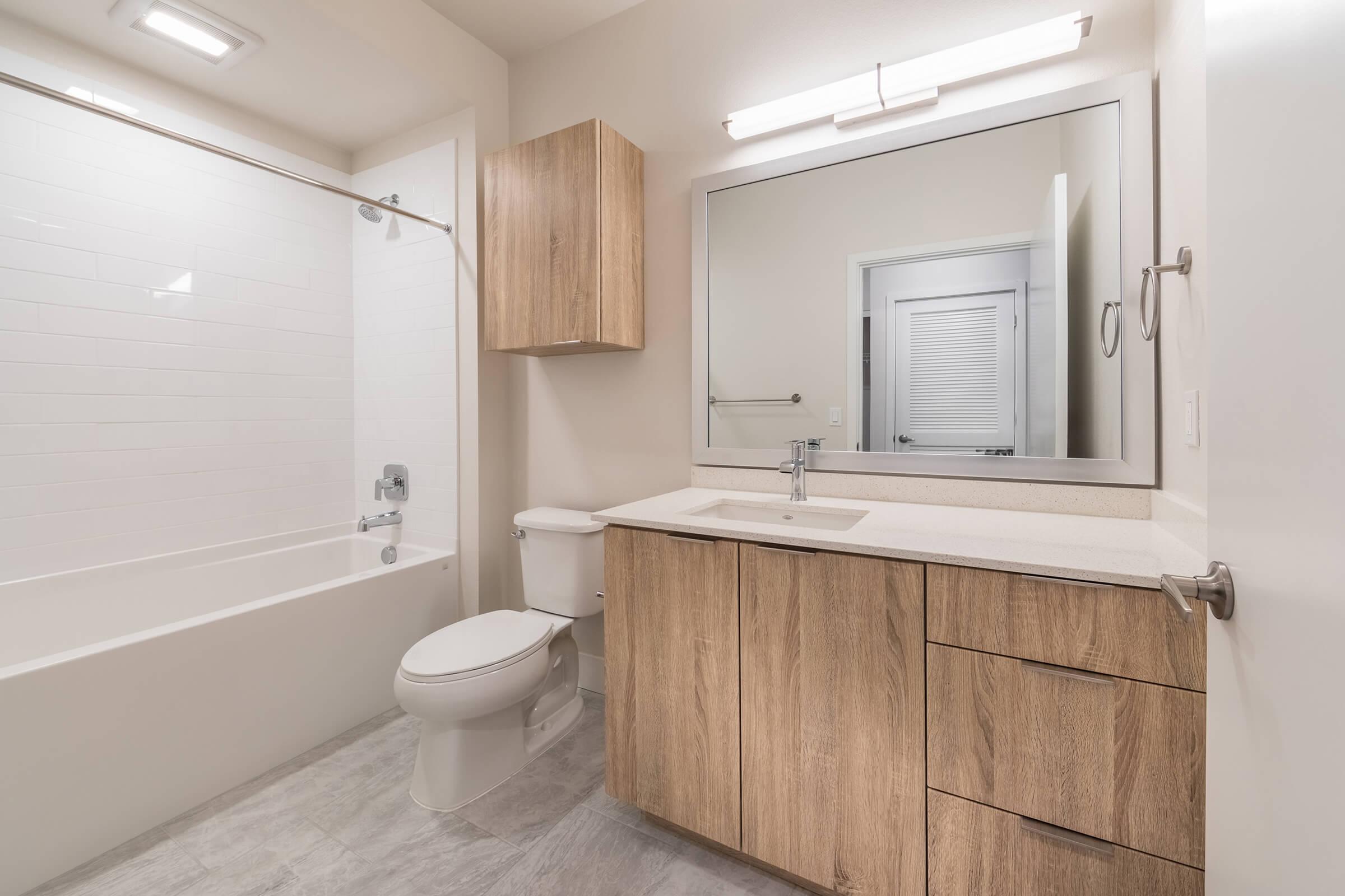 Modern bathroom featuring a bathtub with a shower, a wooden vanity with a sink, a toilet, and a large mirror above the sink. The walls are light-colored and there is soft lighting. The overall design is clean and minimalist, with a neutral color palette.