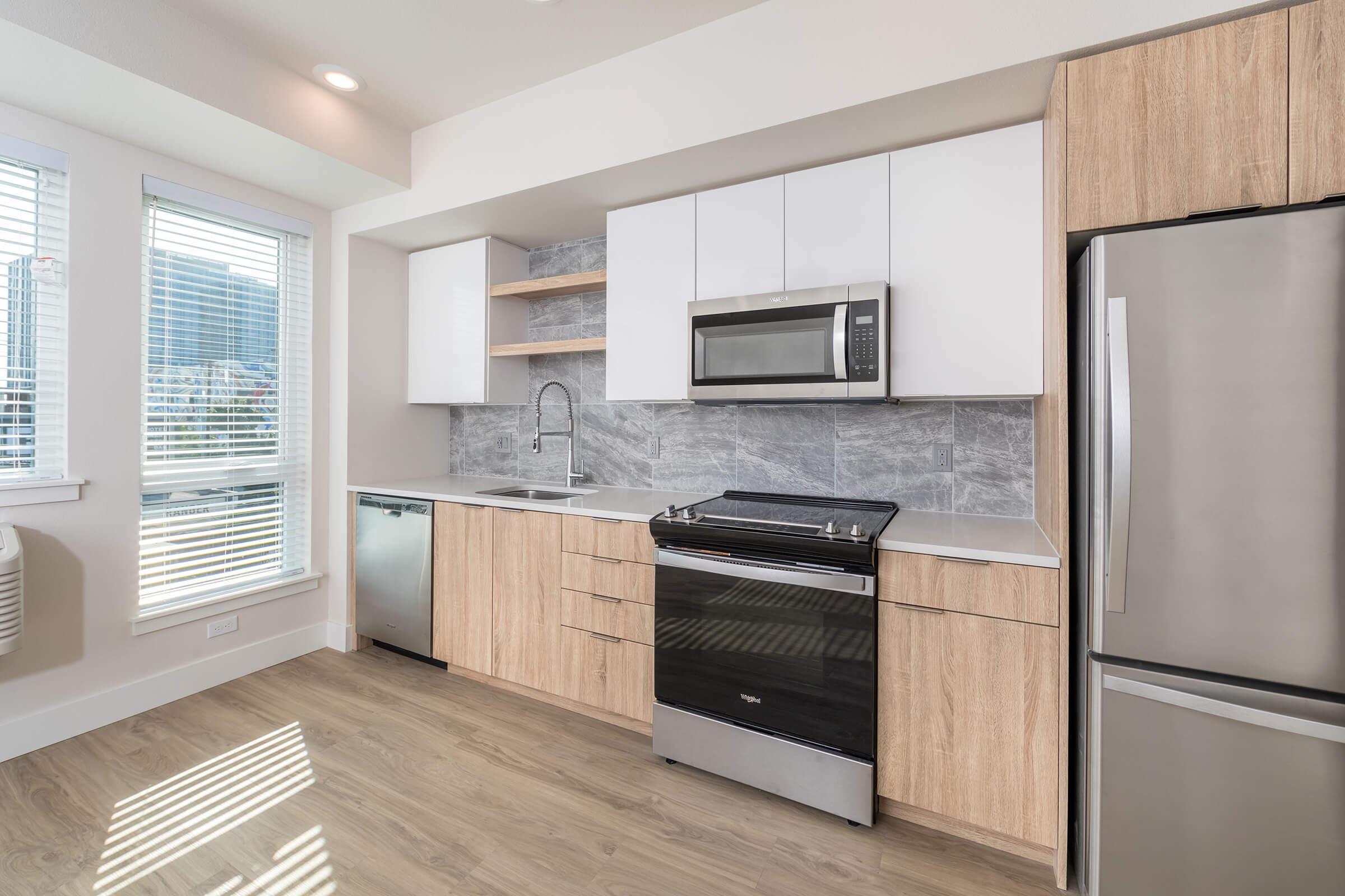 Modern kitchen with white cabinets and wooden accents. Includes a stainless steel refrigerator, microwave, oven, and dishwasher. The countertops are light-colored, and the space features large windows that allow natural light to fill the room. The flooring is light wood laminate.