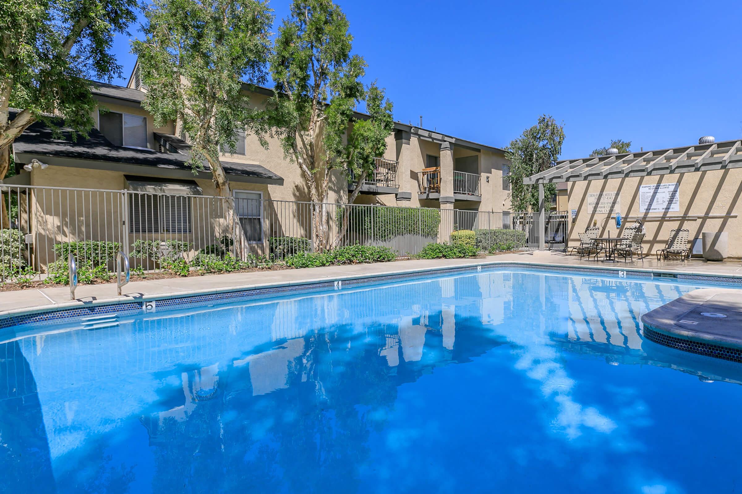 A clear blue swimming pool surrounded by lush greenery and a fenced area. In the background, there are beige apartment buildings and a shaded patio area with tables and chairs. The sky is bright blue, creating a sunny atmosphere.