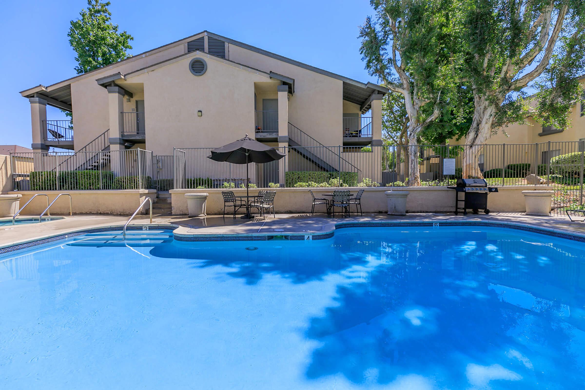 A sparkling blue swimming pool surrounded by lounge chairs and tables, with a shaded umbrella nearby. In the background, there are two-story residential buildings with balconies, framed by lush greenery and clear blue skies. The scene evokes a relaxing, resort-like atmosphere.