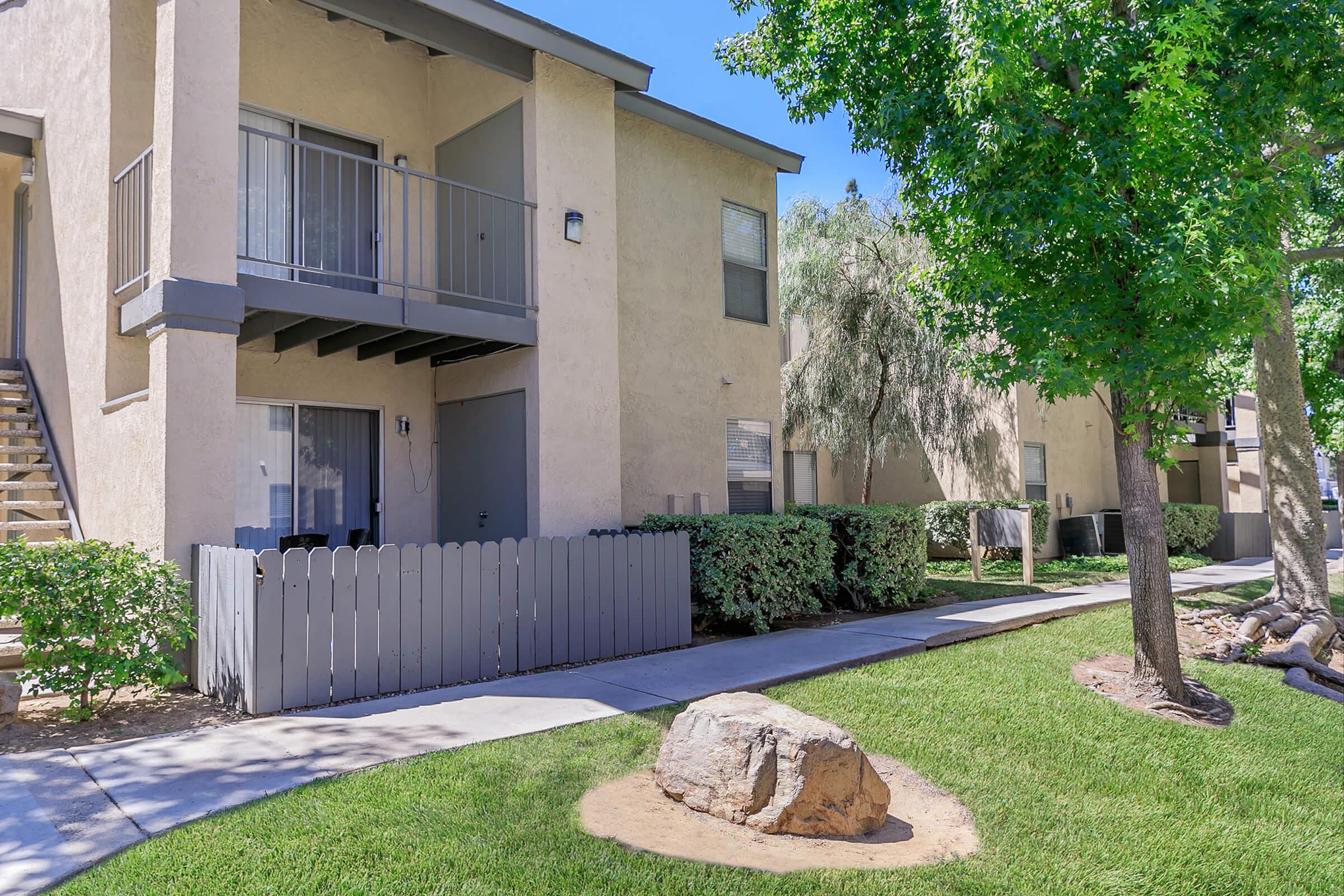 View of a residential apartment complex featuring two-story buildings. The foreground includes a well-maintained lawn with a rock, and a pathway leading to the buildings. There are patio spaces with railings on the upper level, and trees providing shade in the area.