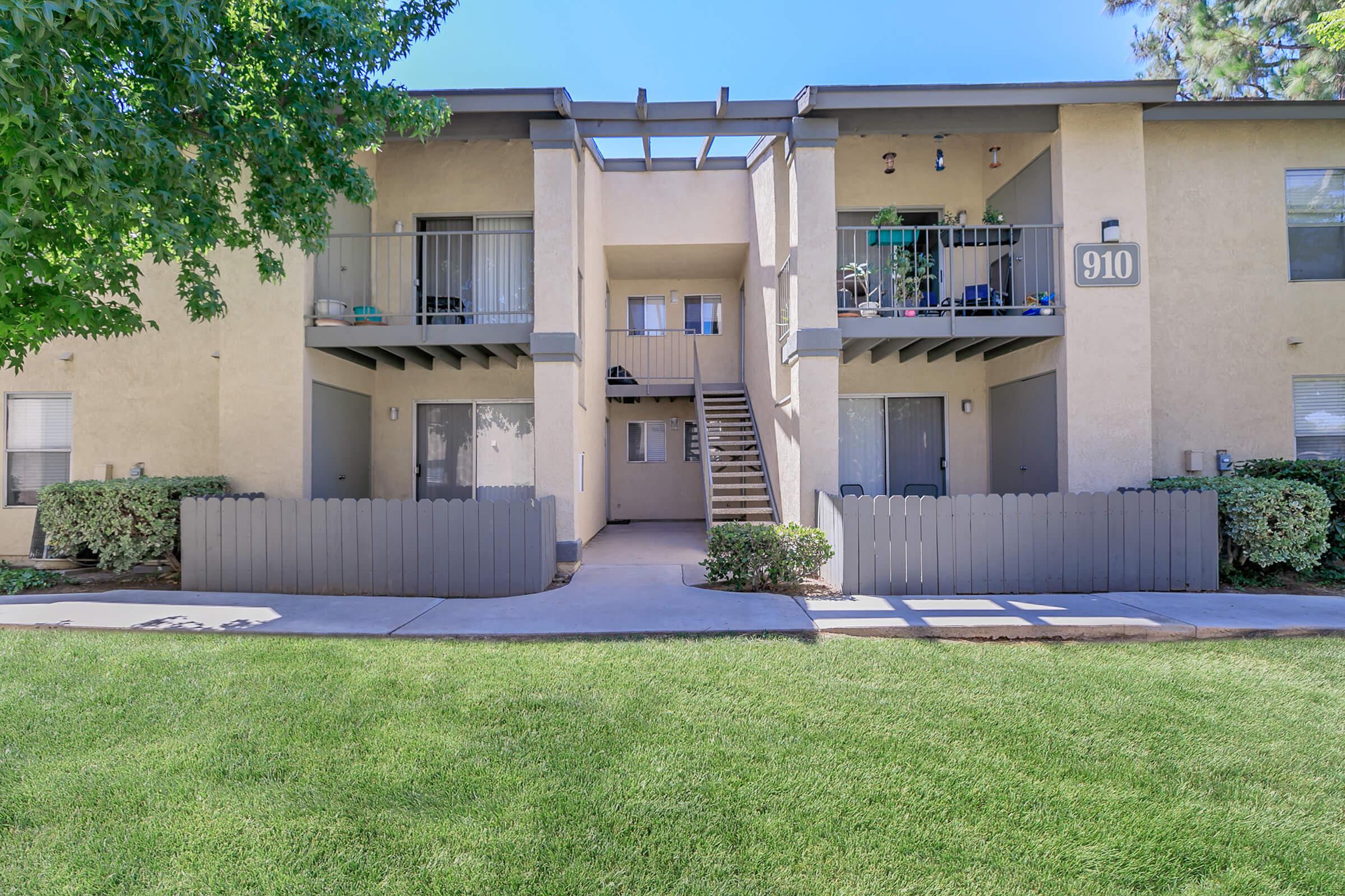 Two-story apartment building with balconies, gray railings, and a central staircase. The building features light-colored walls and small hedges in front of each unit. A well-maintained lawn is visible in front, and the address "910" is displayed on the right side of the entrance. Clear blue sky overhead.