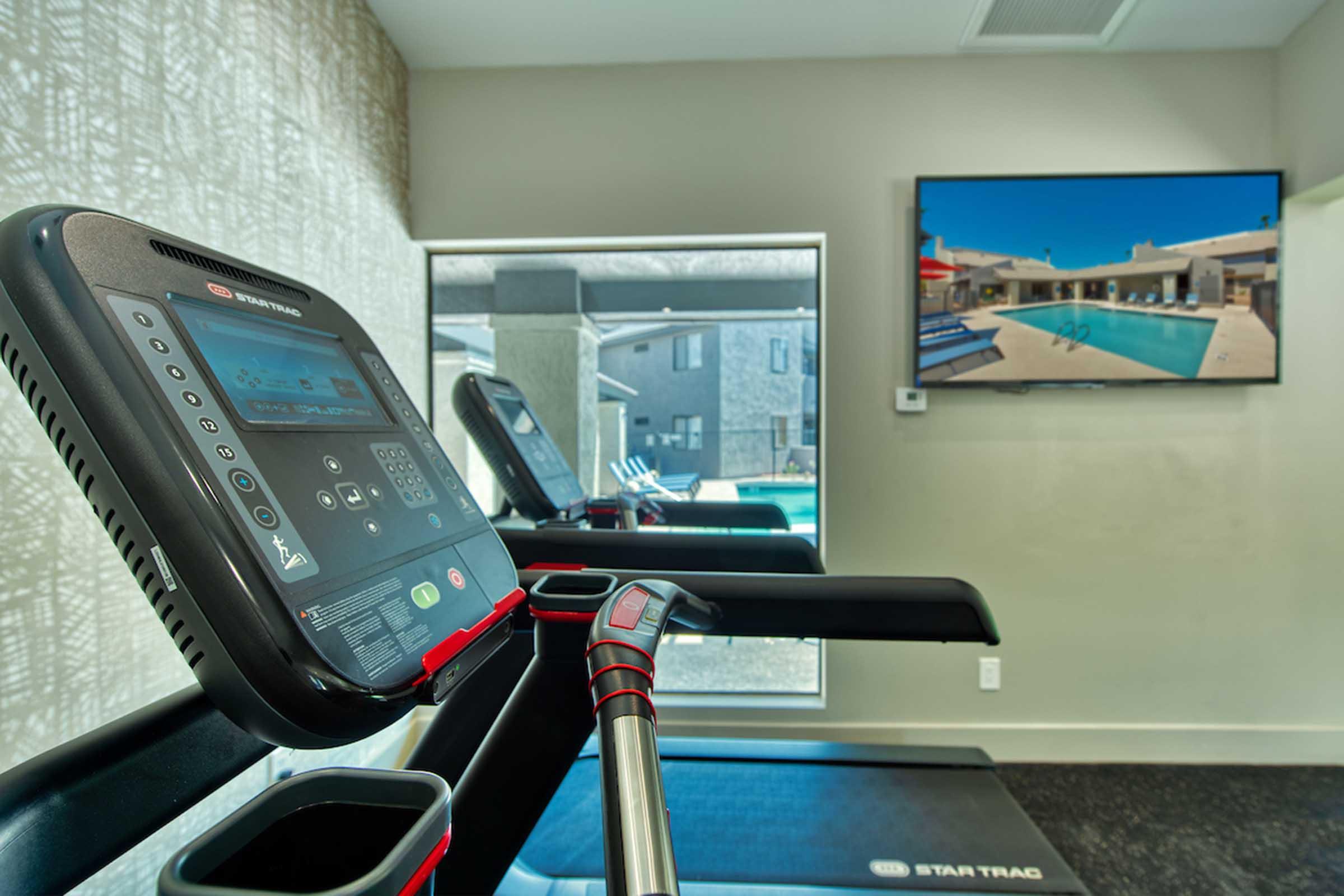 A modern gym with a treadmill in the foreground featuring a digital display. In the background, a large TV screen shows a serene swimming pool scene. Natural light enters through a window, creating a bright and inviting atmosphere.